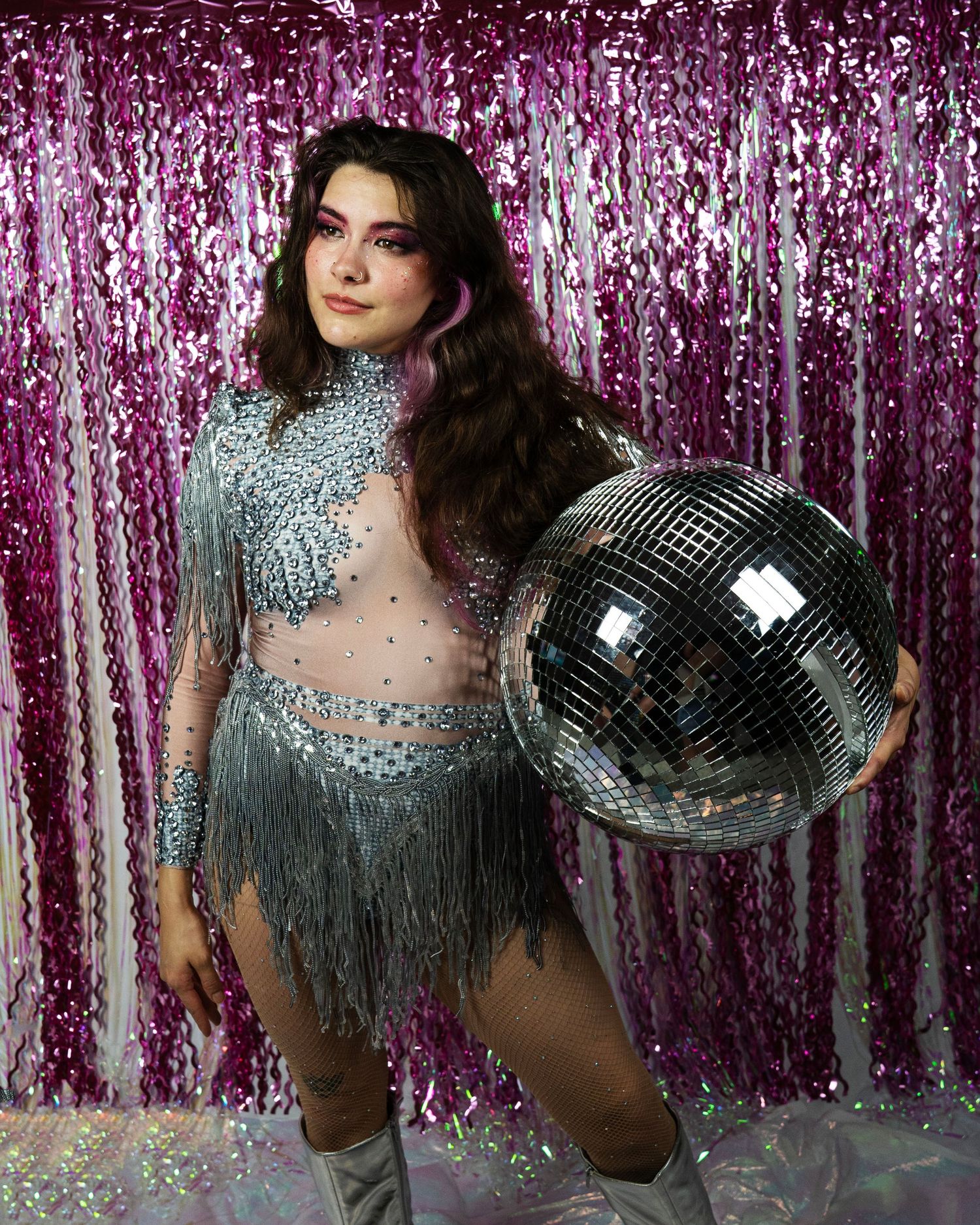 Woman in silver sequin fringe costume holds disco ball against pink tinsel backdrop.