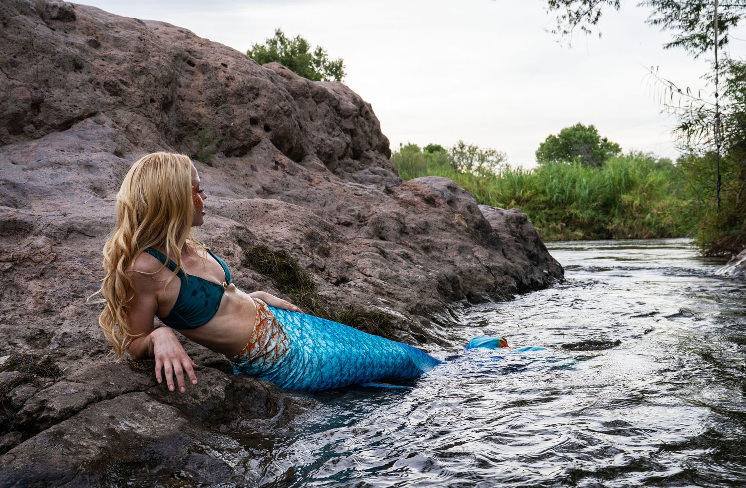 A blonde woman wearing a blue mermaid tail sits on rocks beside a gently flowing river. Fantasy Photoshoot