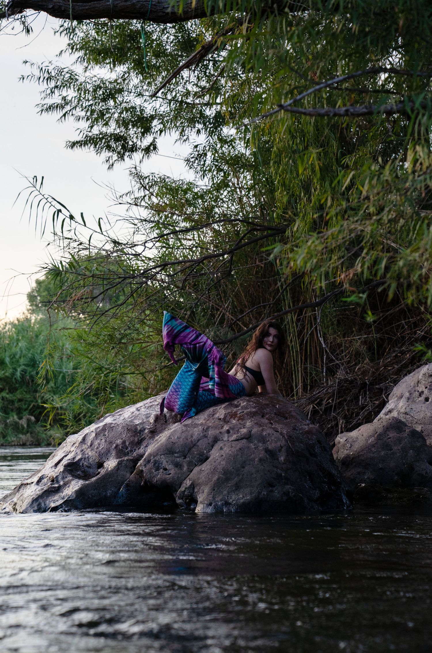 A woman wearing a vibrant mermaid tail costume poses on riverside rocks surrounded by green foliage. Mermaid photoshoot, fantasy photoshoot.