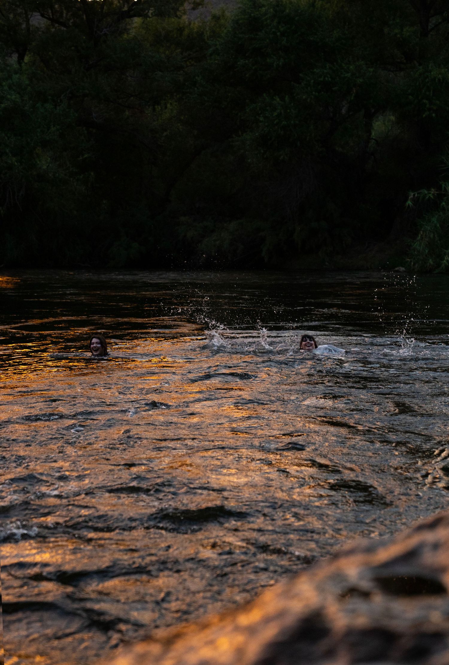 Two people swimming in golden-lit river at dusk with splashing water. Fantasy photoshoot. Arizona photographer.