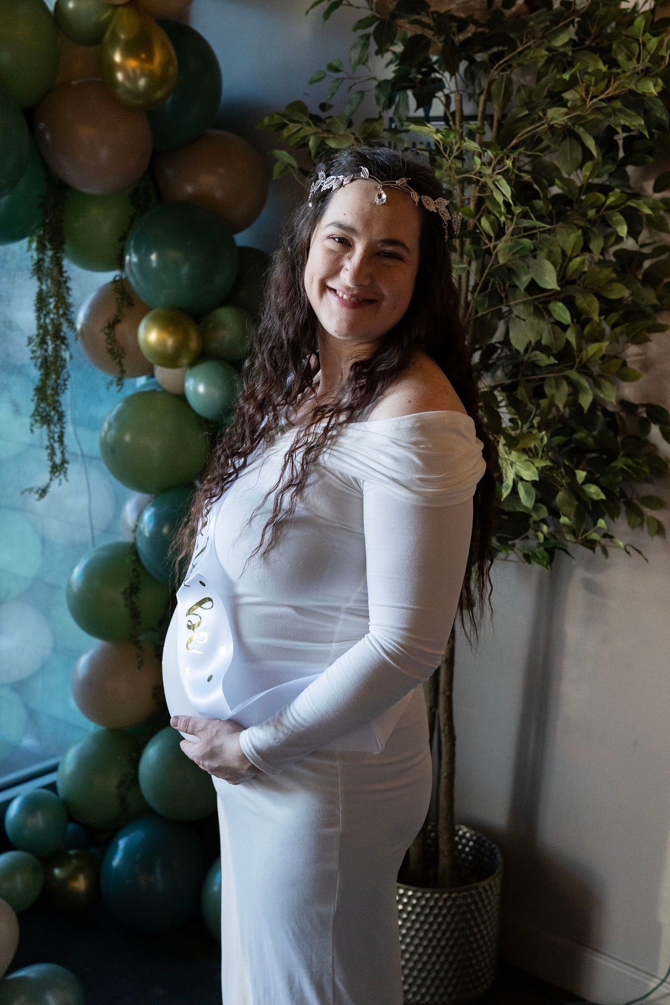 Pregnant woman in white off-shoulder dress with floral crown smiles at baby shower with green balloon arch.