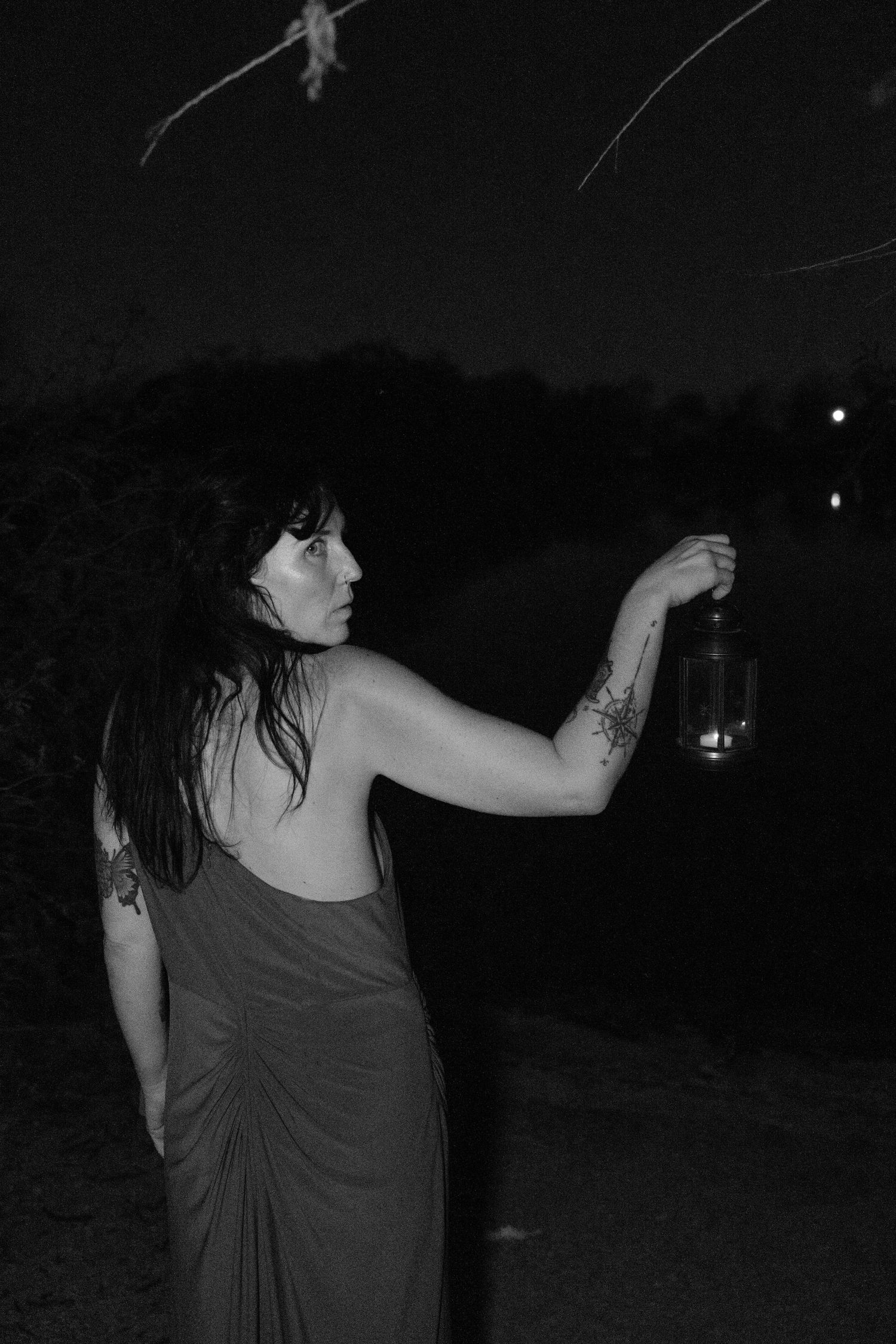 Black and white photo of a woman in a draped dress holding a lantern at night beside a dark lake, her arm tattoos visible.