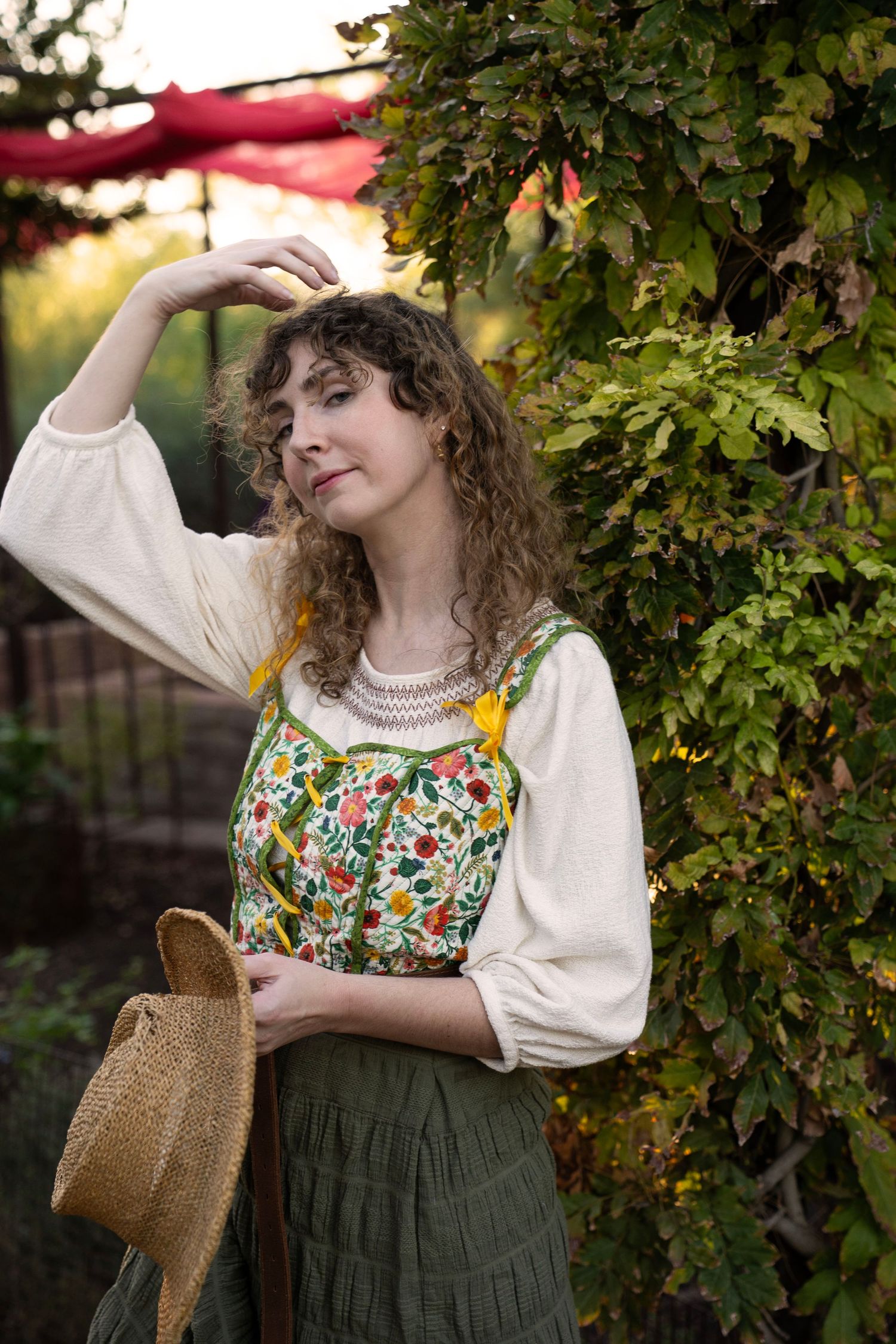 Woman with curly hair in a floral embroidered vest and cream blouse holds a straw hat beside a leafy vine in a sunlit garden.