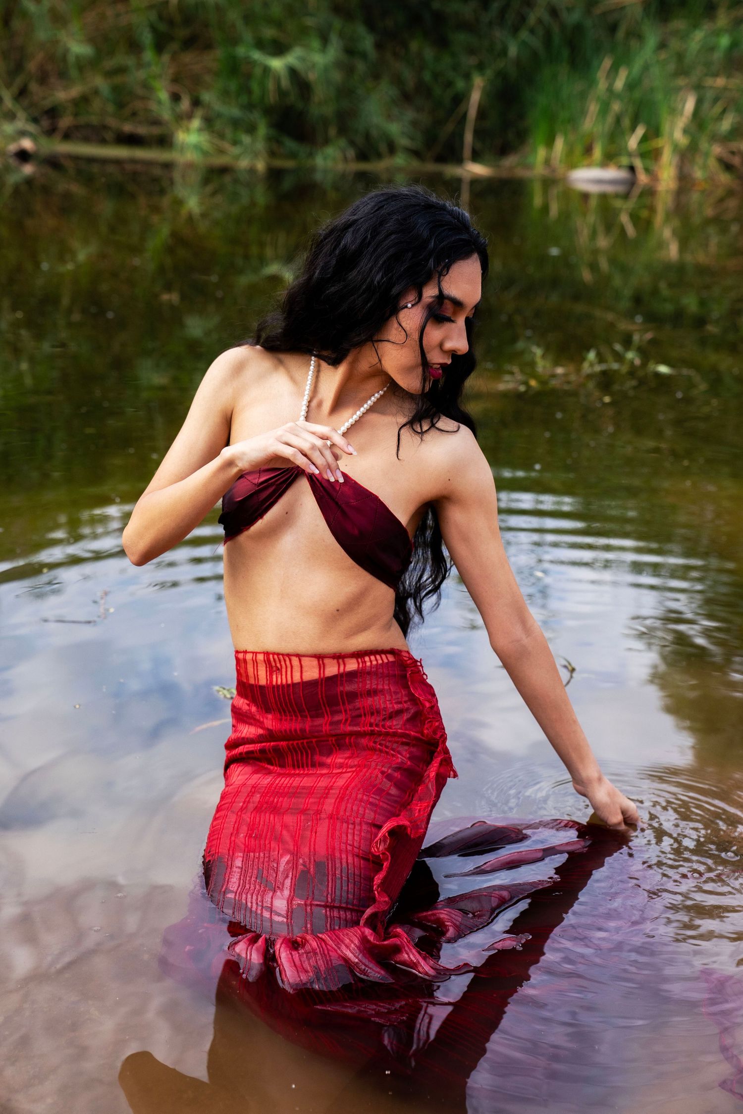 A dark-haired woman in a red skirt and burgundy top wades through a calm river surrounded by lush greenery.