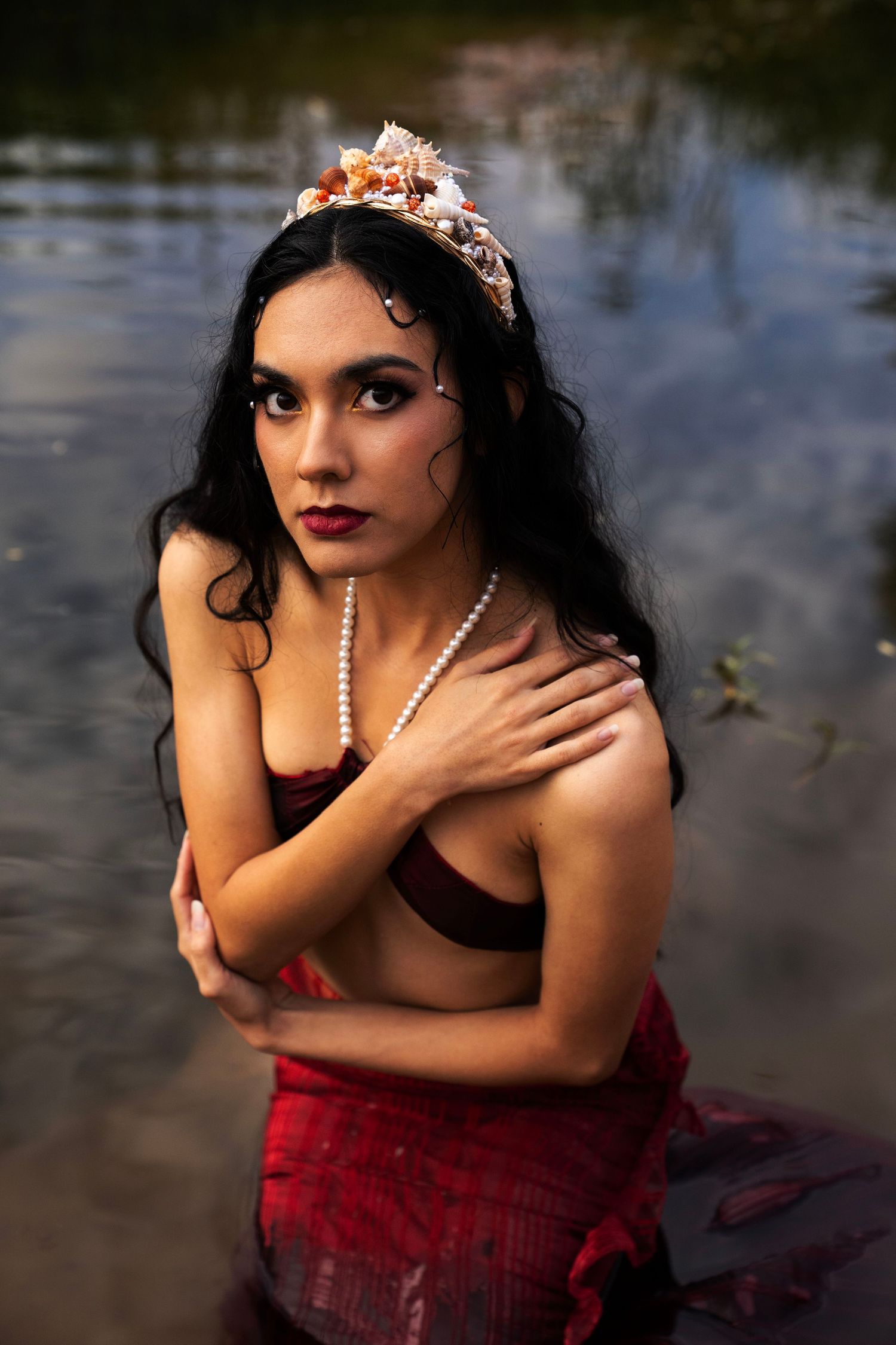 A woman wearing a floral crown and pearl necklace poses in a river, arms crossed, with a dramatic mermaid-inspired look.