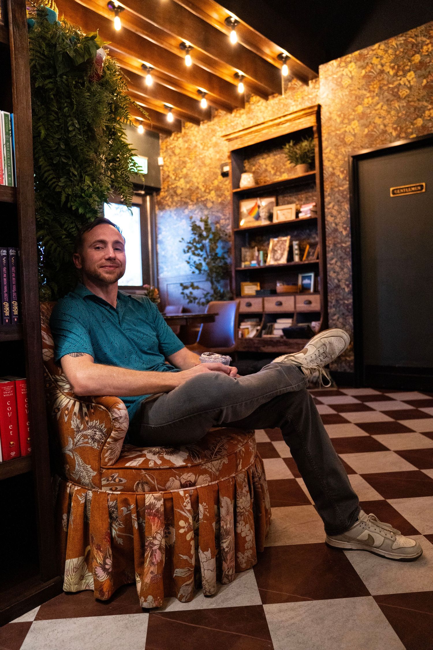 Man in teal shirt relaxing in floral chair in cozy vintage-style lounge with string lights.
