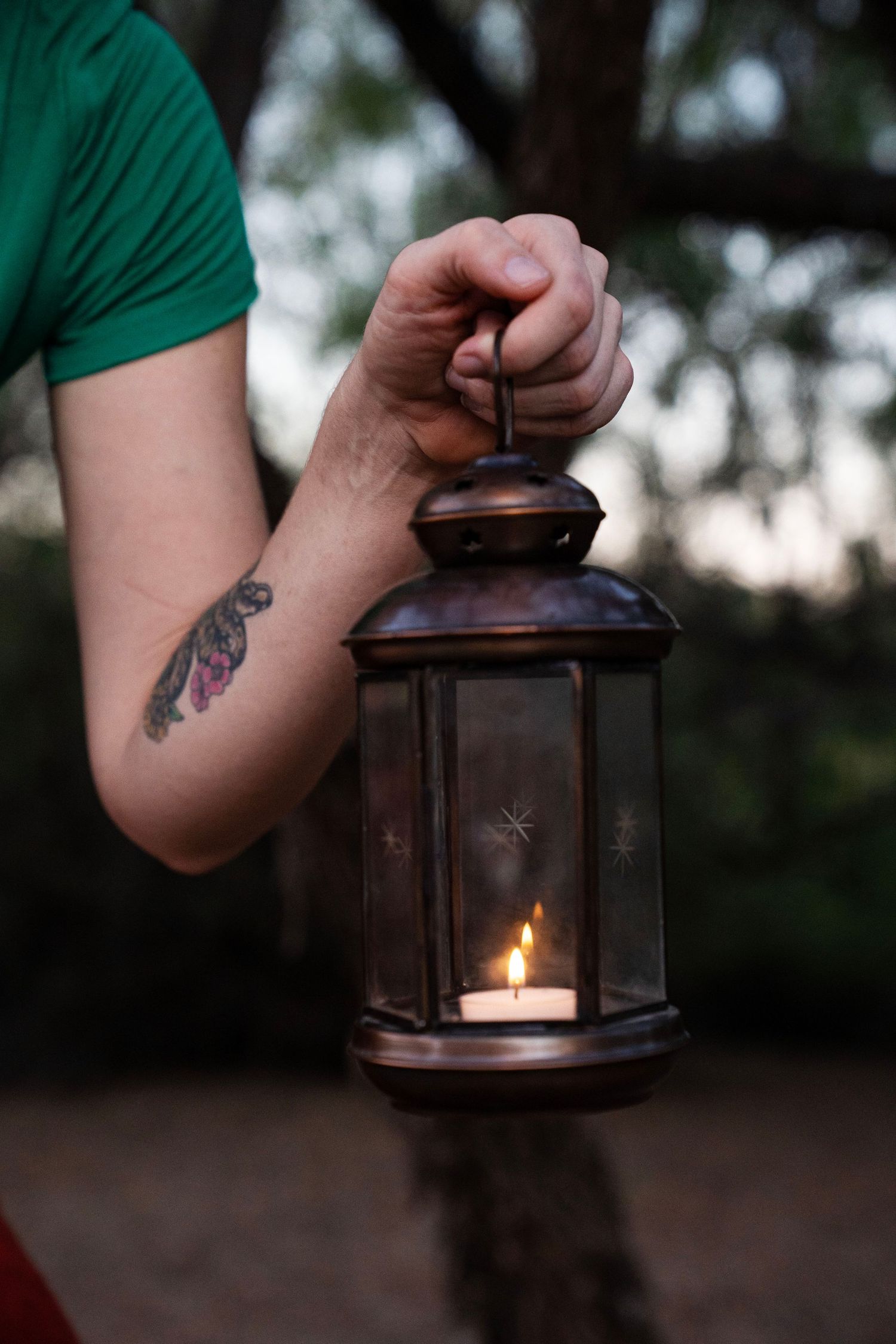 Person in a green shirt with a floral forearm tattoo holds a vintage copper lantern with a lit candle outdoors at dusk.