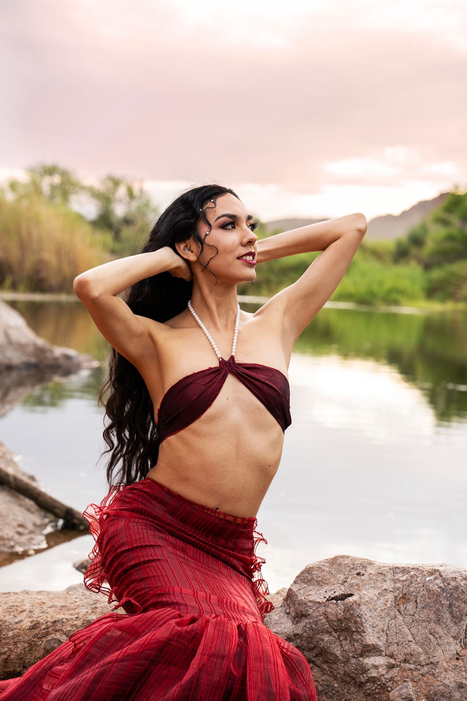 A woman in a red mermaid costume sits on rocks by a river, posing with hands behind her head at sunset.
