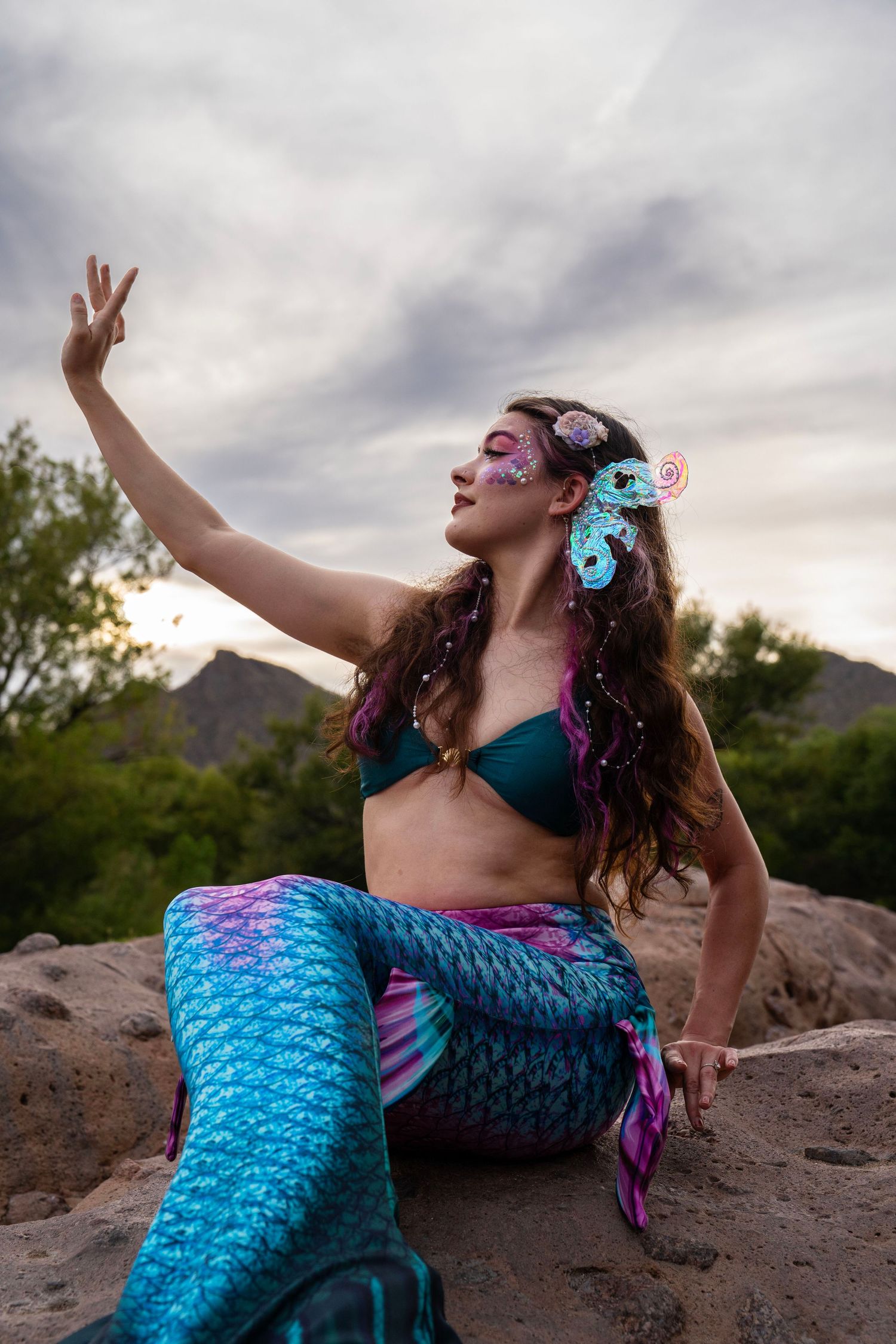 Woman in a blue and pink mermaid costume with face paint and seahorse hair accessories posing on rocks. Fantasy Photoshoot