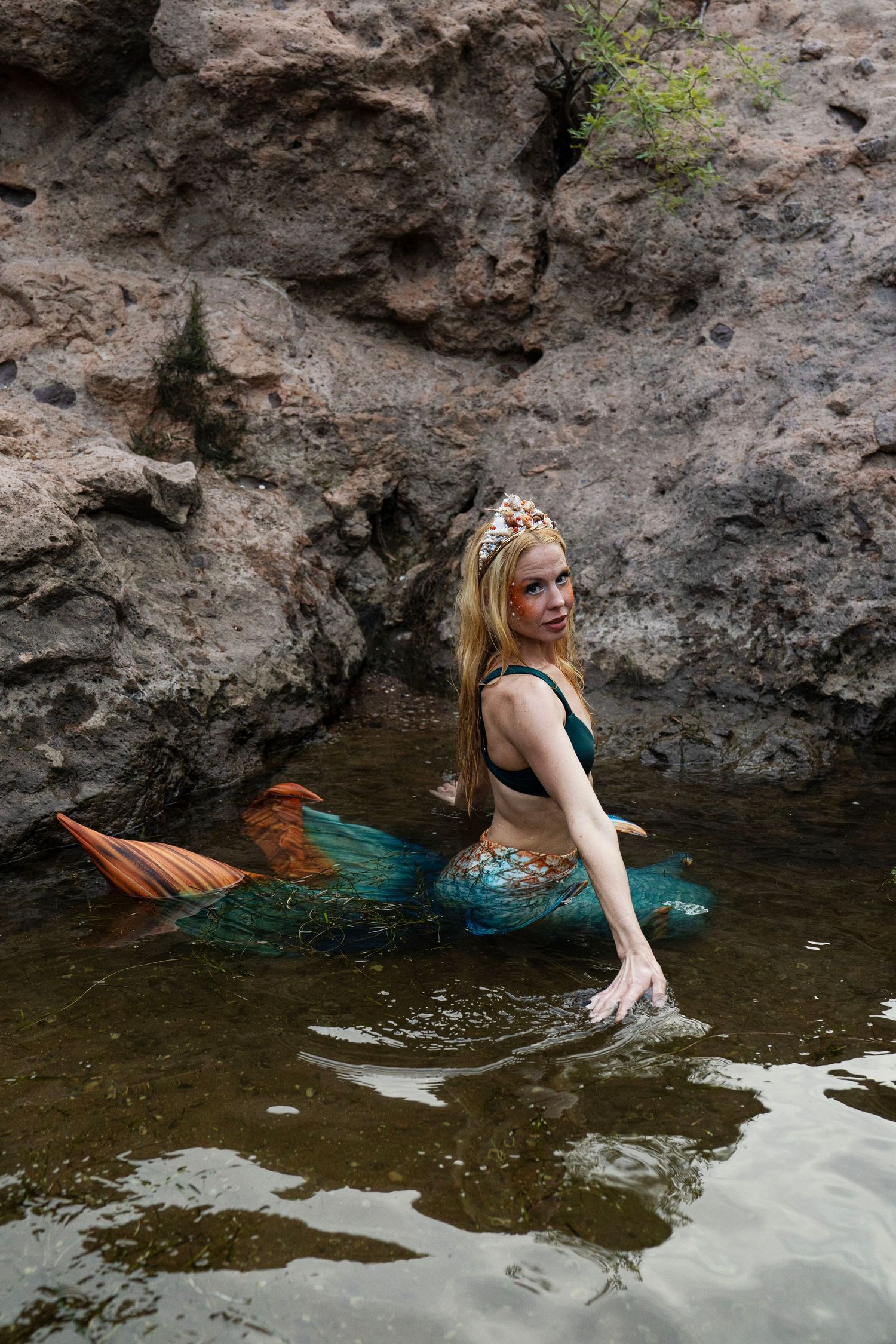 Woman in mermaid cosplay with colorful teal and orange tail sitting in shallow rocky water. Fantasy Photoshoot