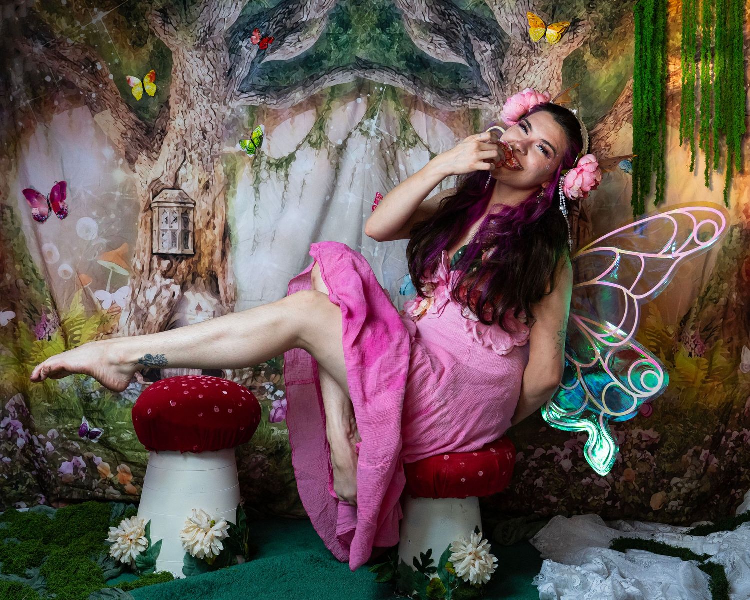 Woman dressed as fairy in pink dress with colorful wings poses on red mushroom in magical forest backdrop. Fantasy Photoshoot