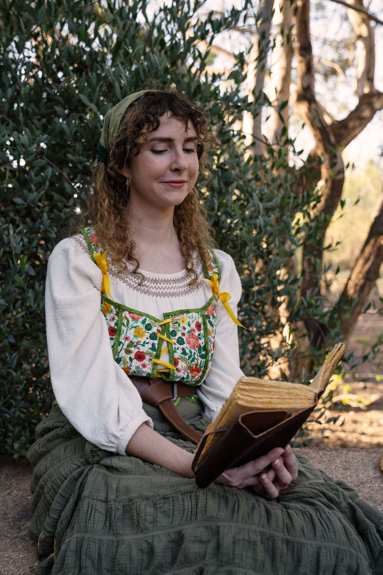 Woman with curly hair and a green headscarf reads a leather-bound book outdoors, wearing a floral bodice, cream blouse and green skirt.