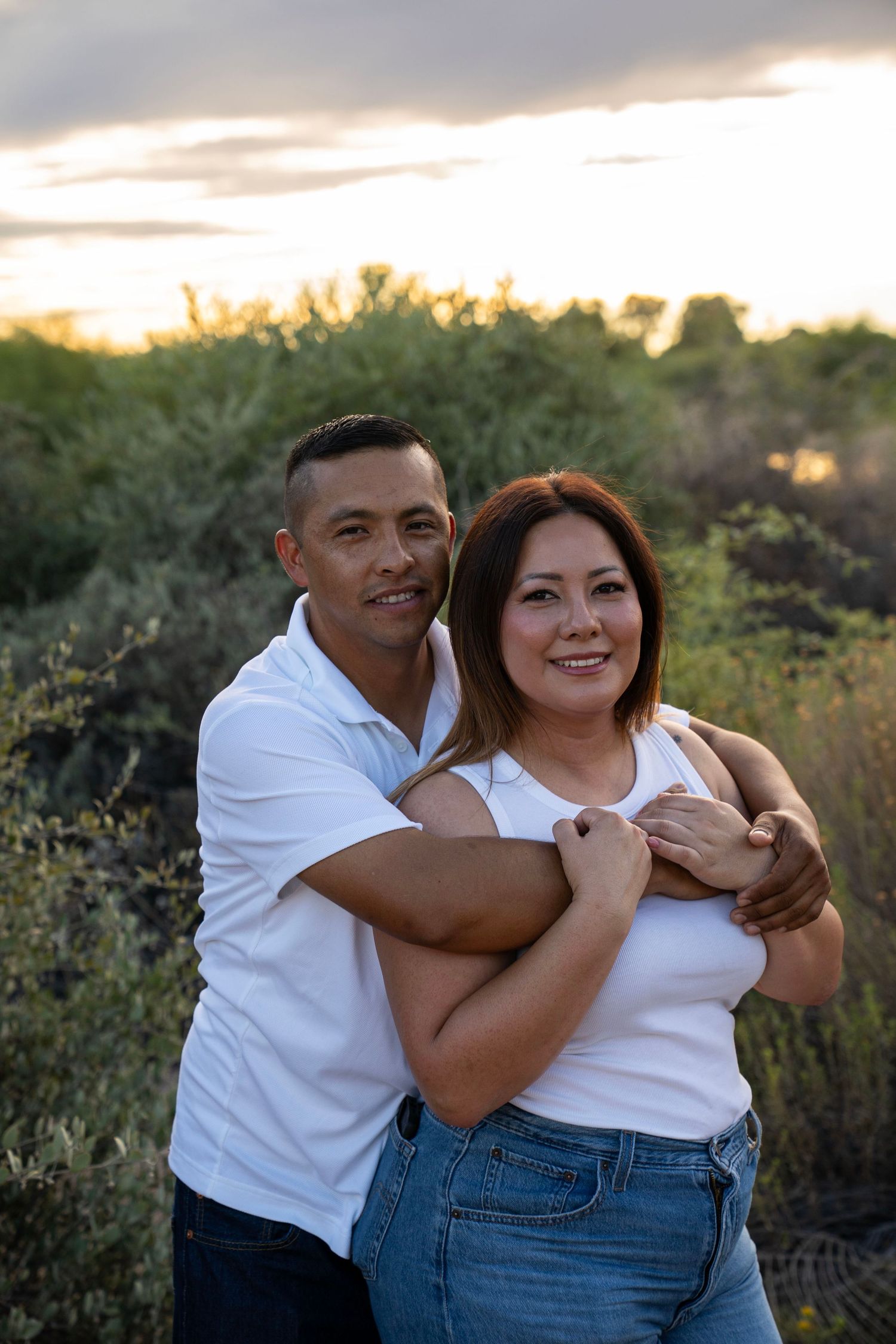 A couple embraces outdoors at sunset, both wearing white shirts and jeans, smiling warmly among desert greenery.