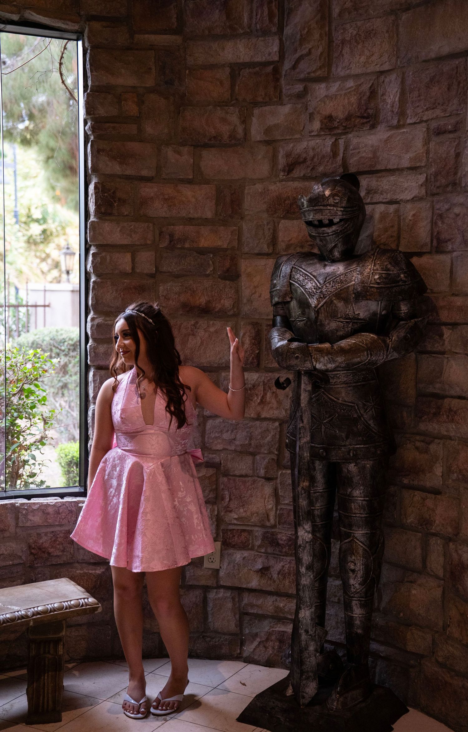 Young woman in pink dress poses beside medieval knight armor statue in stone castle interior near window.