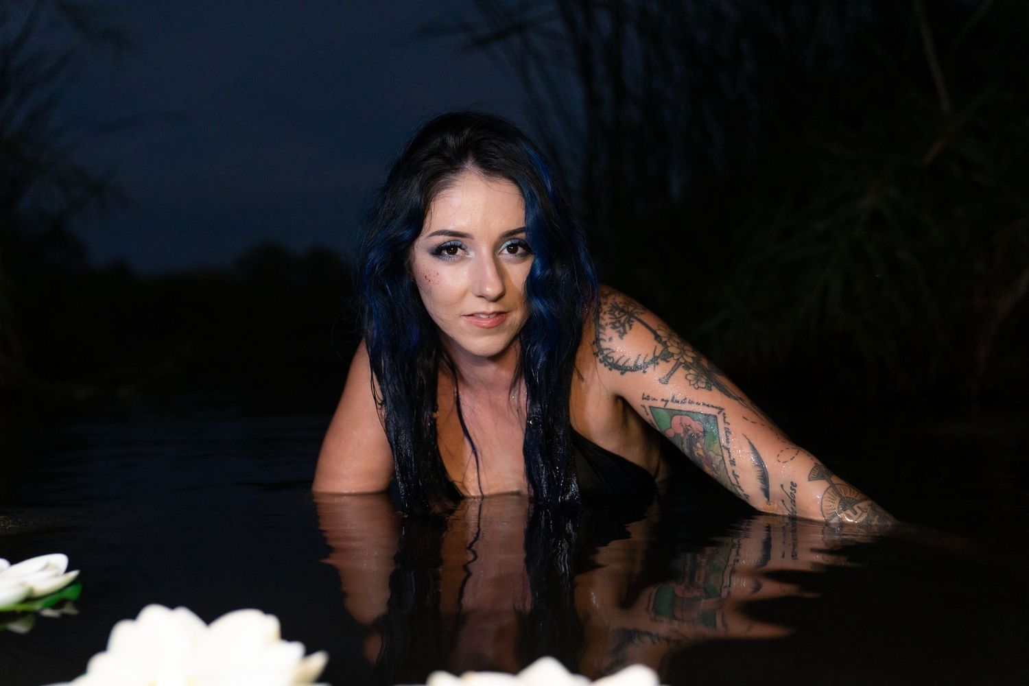 Tattooed woman with blue-black hair emerges from dark water surrounded by white flowers at night.