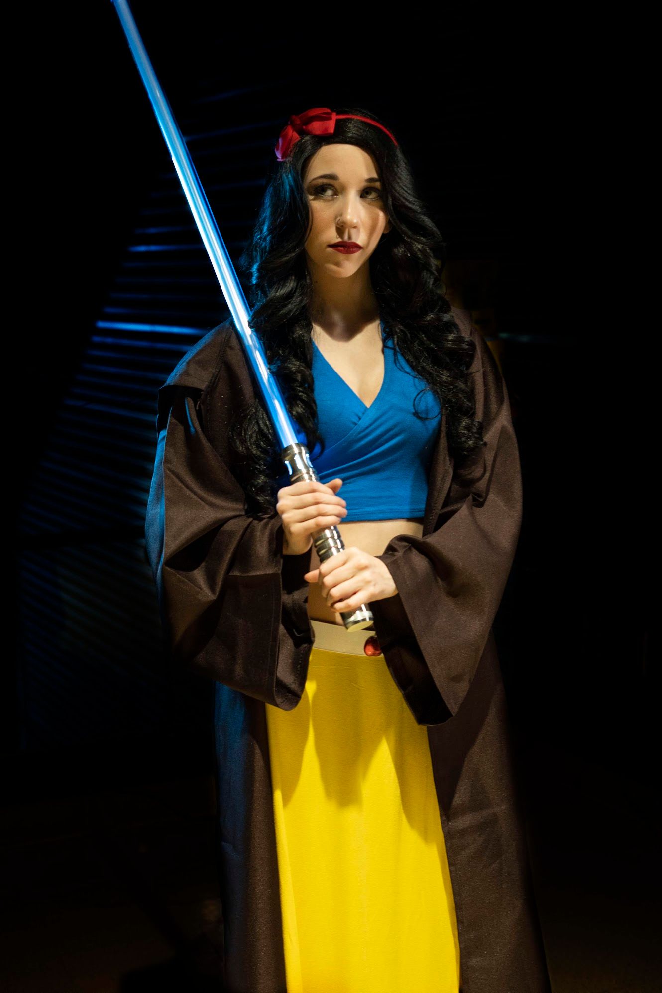 Cosplayer in a Snow White-inspired outfit with red bow, blue top and yellow skirt, wearing a Jedi robe and holding a blue lightsaber.