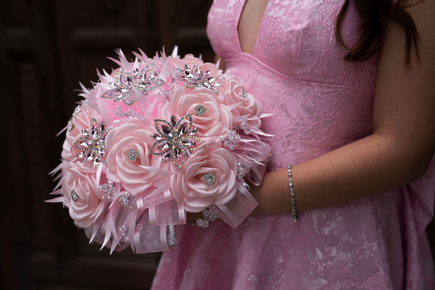 Close-up of elegant pink bouquet with satin roses, feathers, and crystal brooches held by woman in pink dress.
