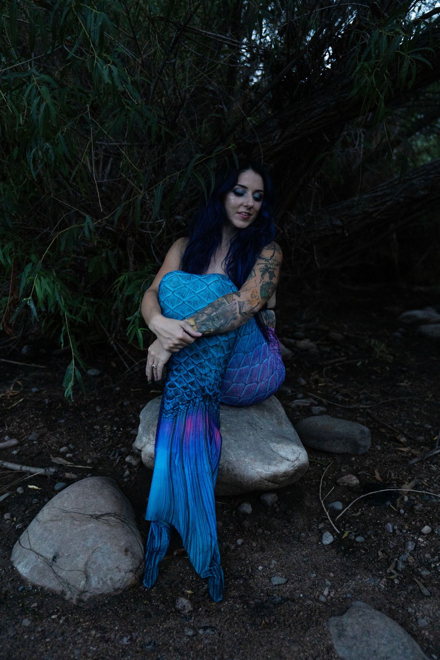 Tattooed woman in blue mermaid costume sitting on rocks in a dark forest setting.