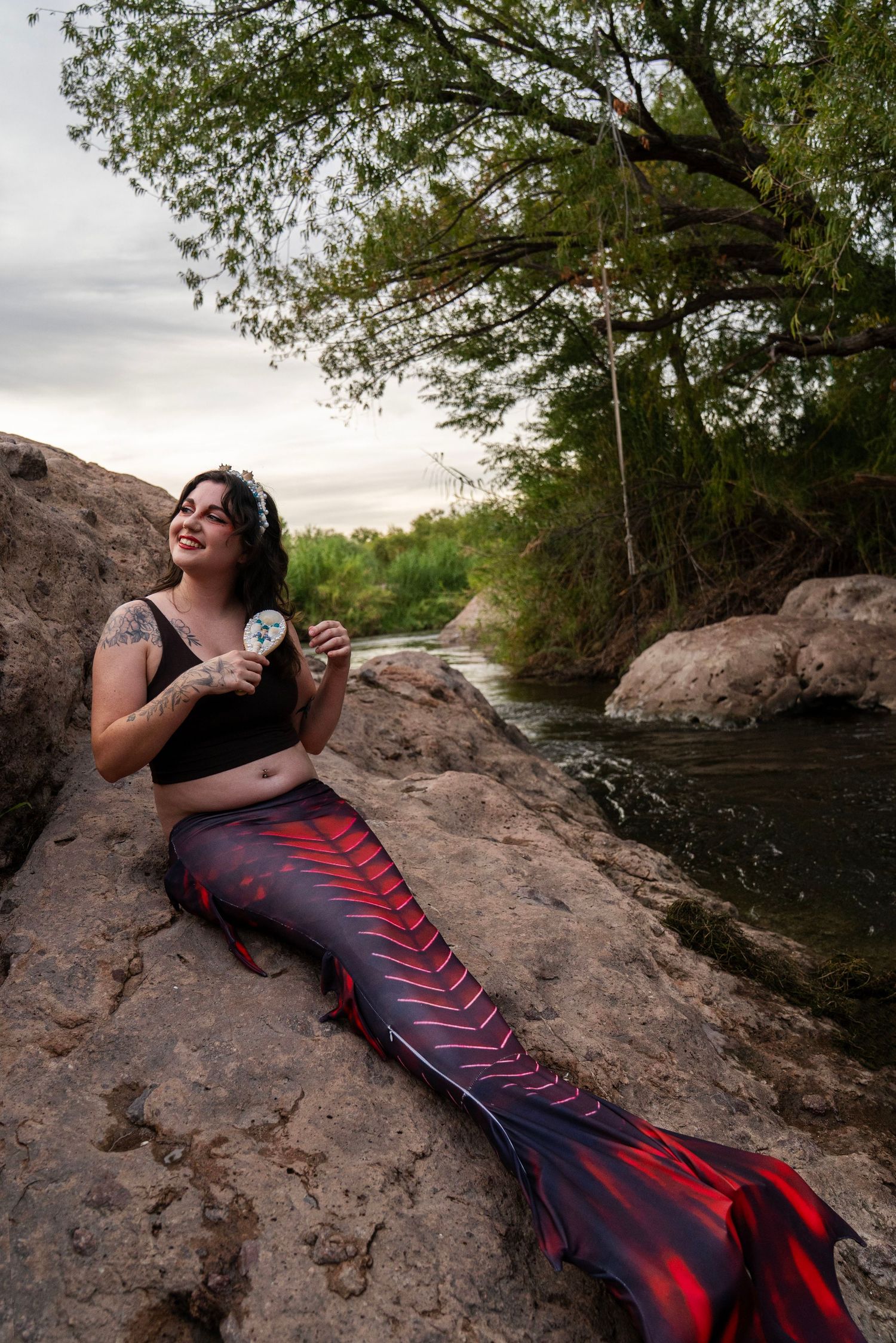 A smiling woman in a red mermaid tail costume sits on rocks near a river. Fantasy Photoshoot