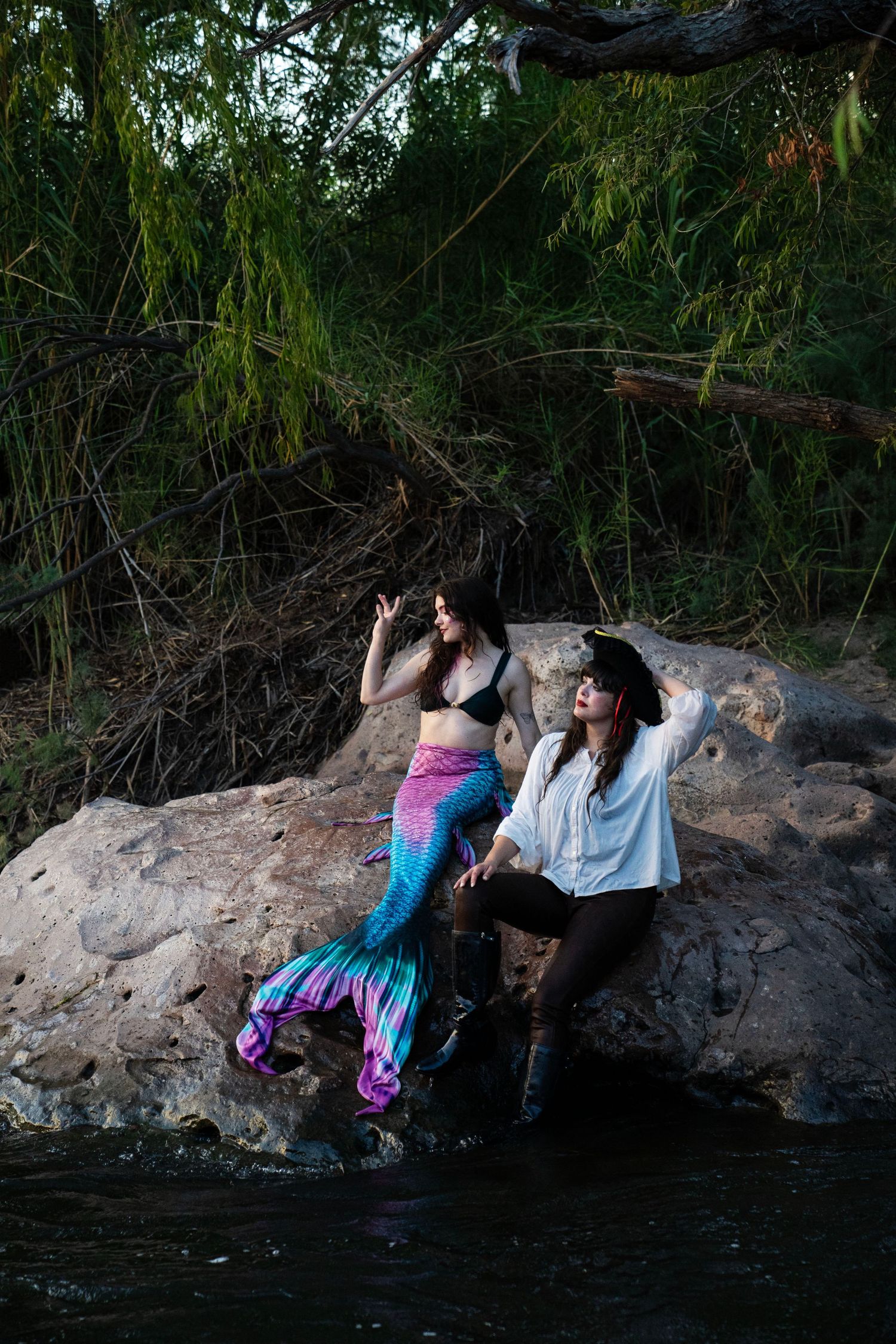 A woman in a mermaid costume and a person in a pirate outfit sit together on riverside rocks. Mermaid photoshoot, fantasy photoshoot.
