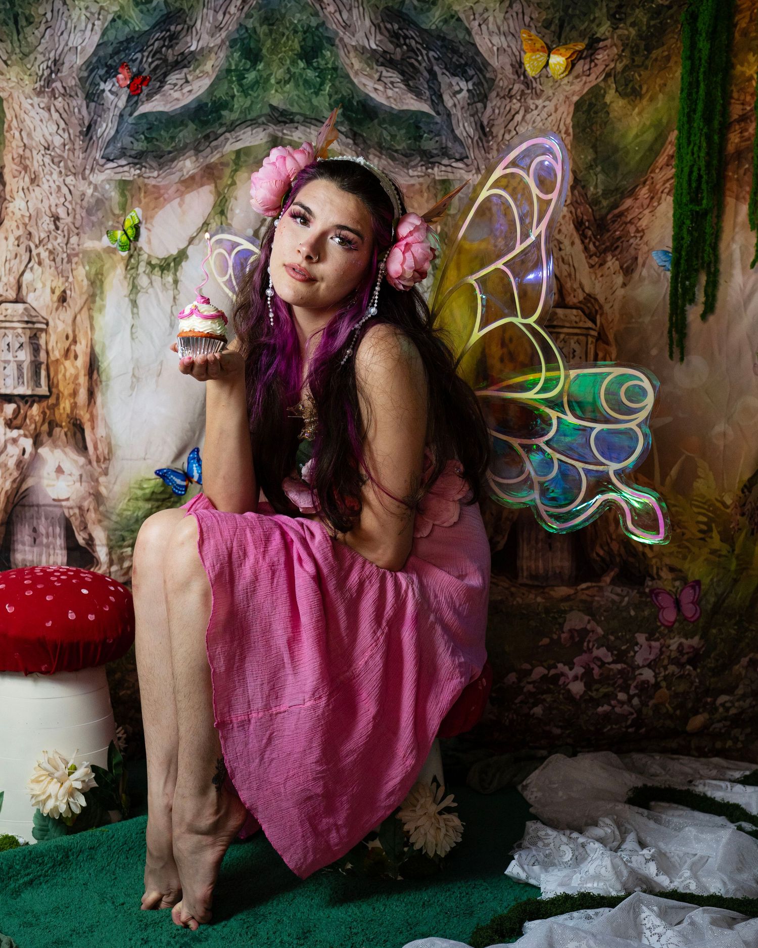 Young woman in pink fairy dress with iridescent wings holds cupcake by red mushroom in forest set. Fantasy Photoshoot