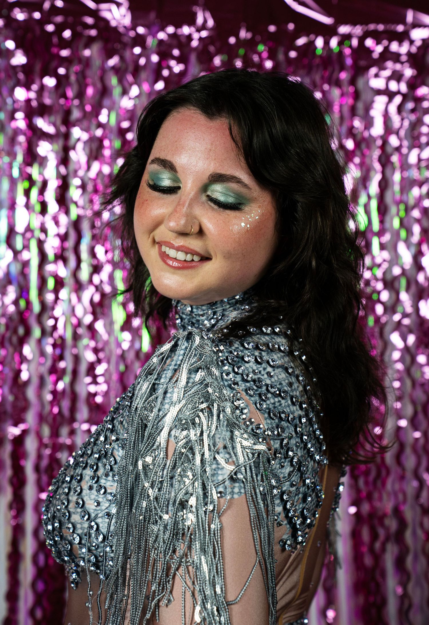 Woman with green glitter eye makeup smiles in silver beaded fringe costume on pink backdrop.