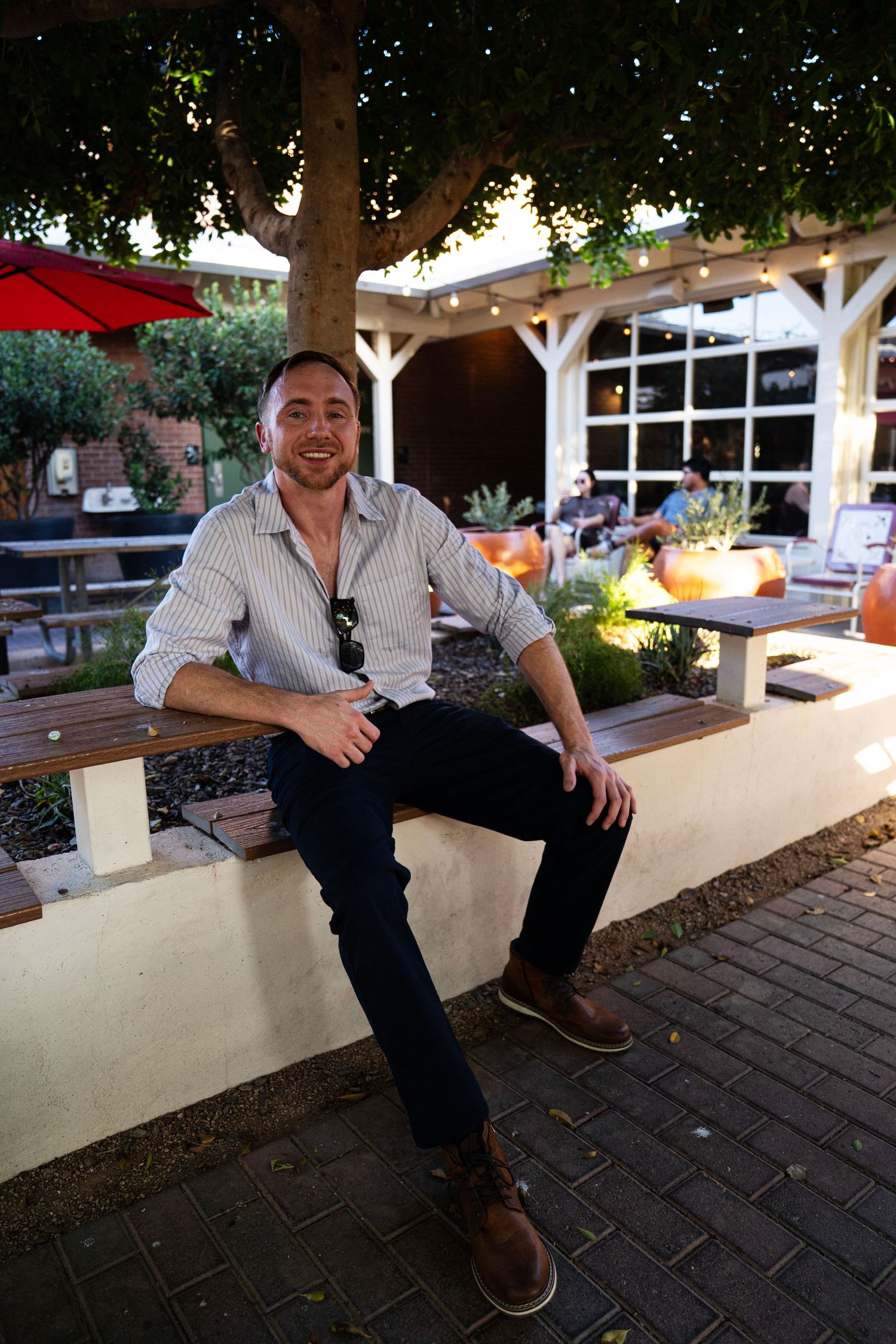 Smiling man sitting on outdoor bench at patio restaurant under tree with string lights at dusk.
