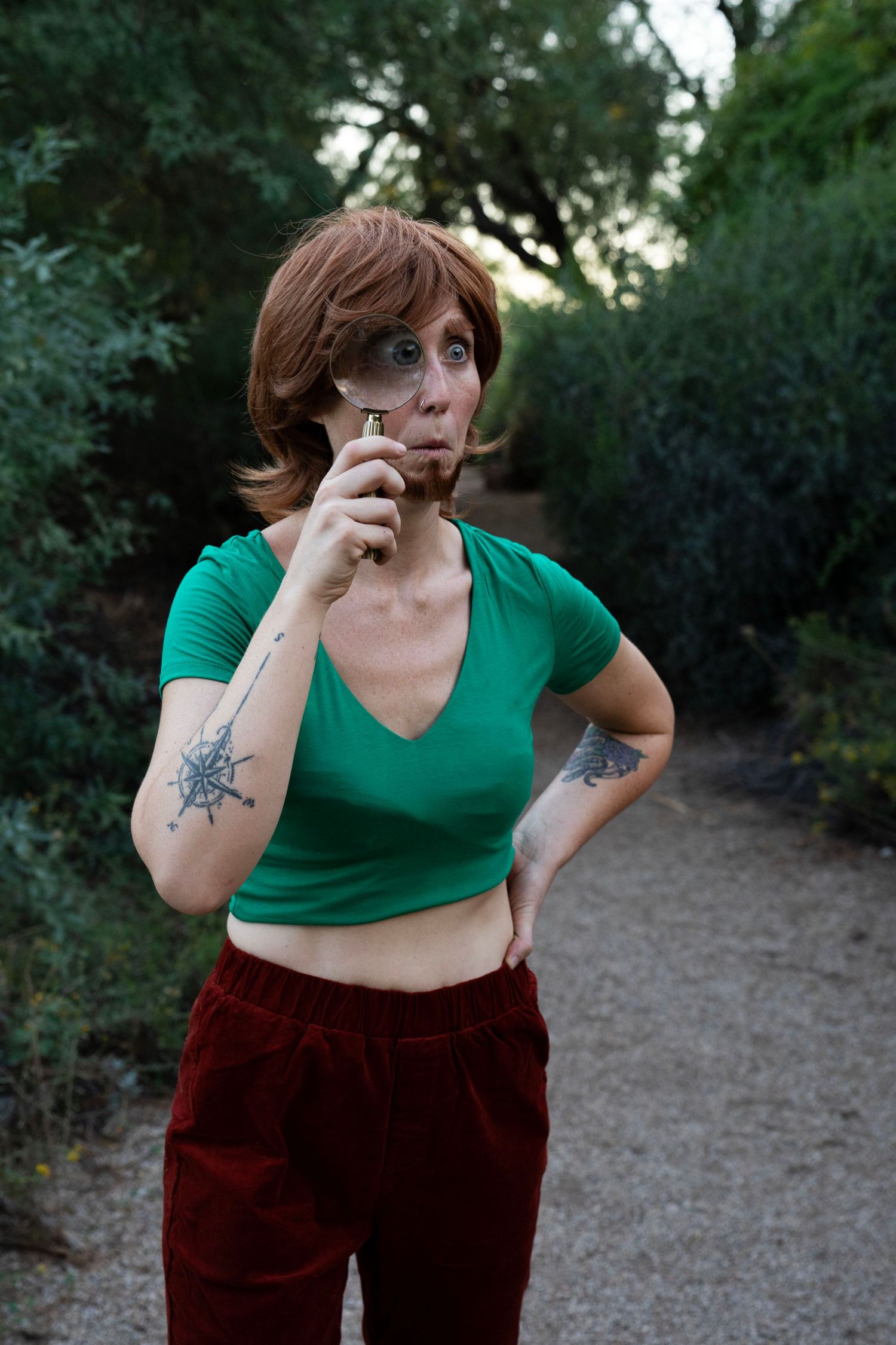 Person with auburn hair and arm tattoos holds a magnifying glass to one eye, wearing a green crop top and red velvet pants on a trail.