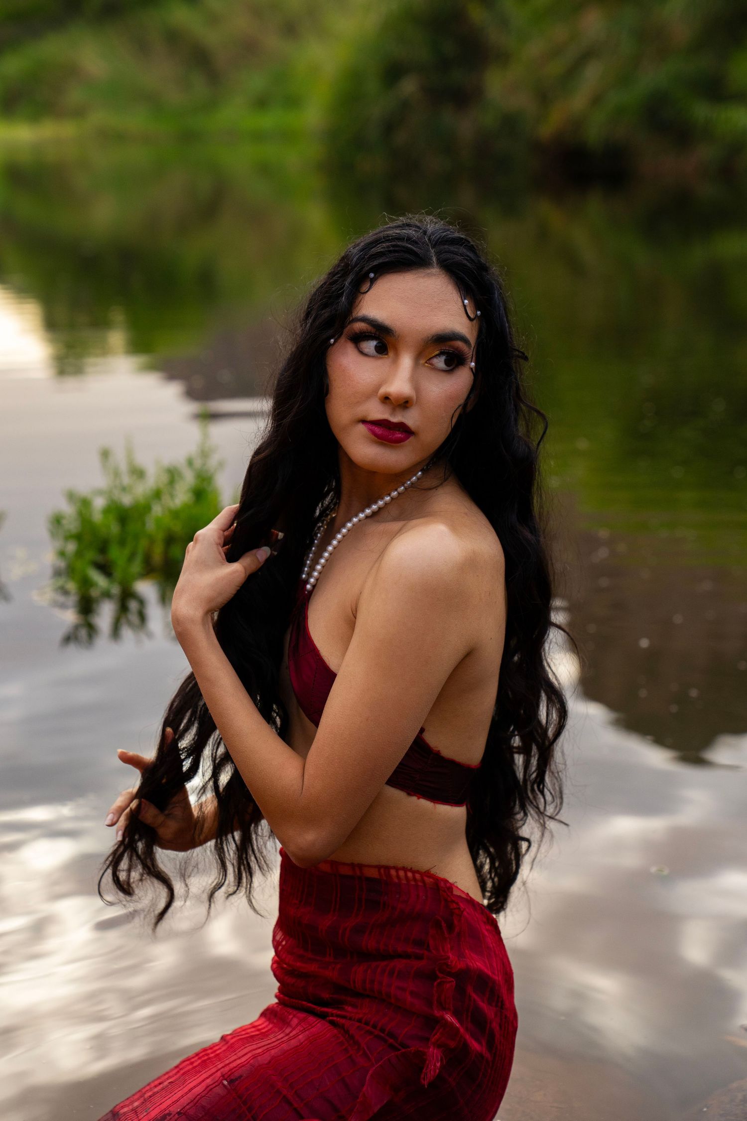 A woman with long black hair and red lips poses in shallow water wearing a pearl necklace and red skirt outdoors.