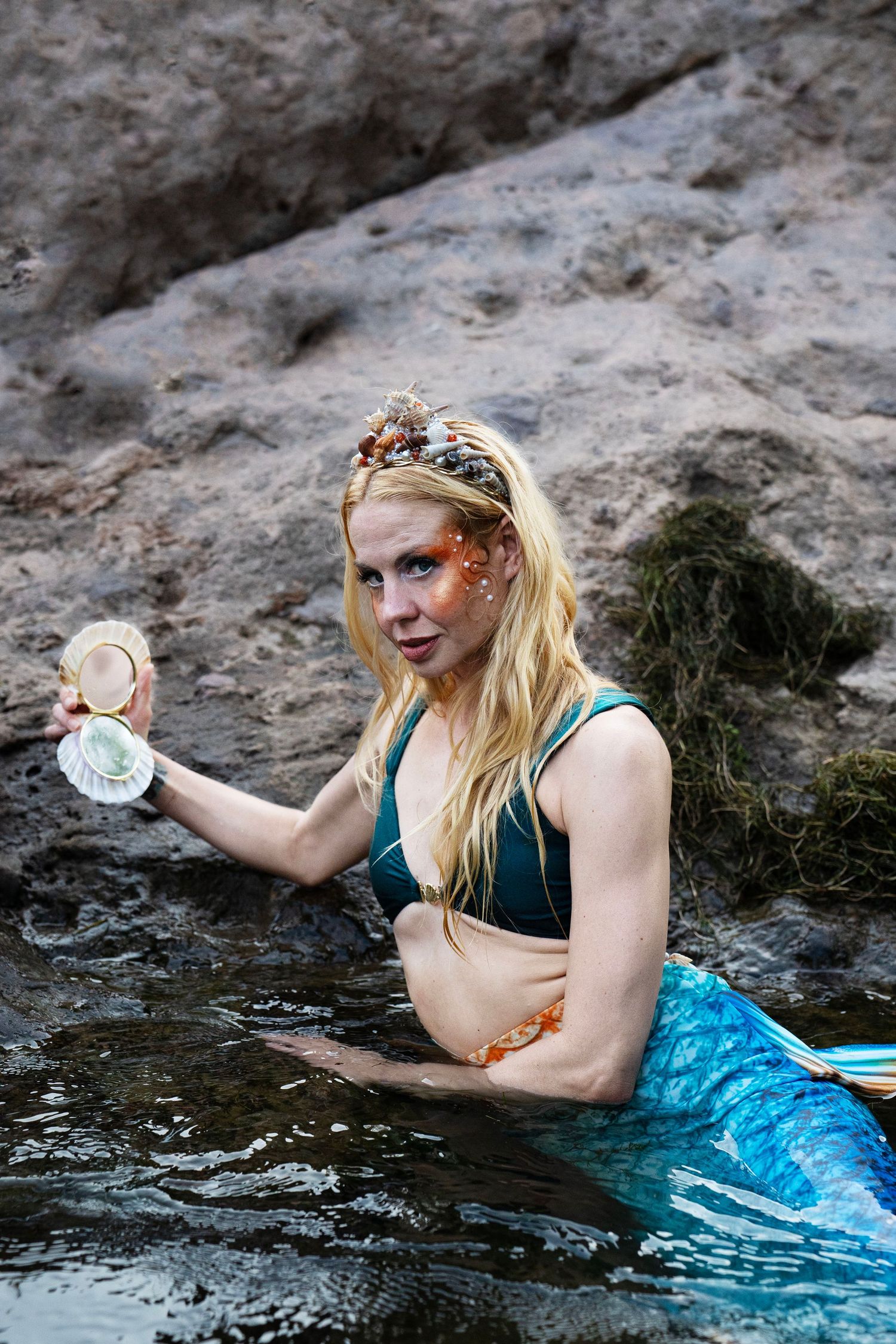A blonde mermaid with a floral crown and glitter makeup holds a seashell while sitting in water. Fantasy Photoshoot