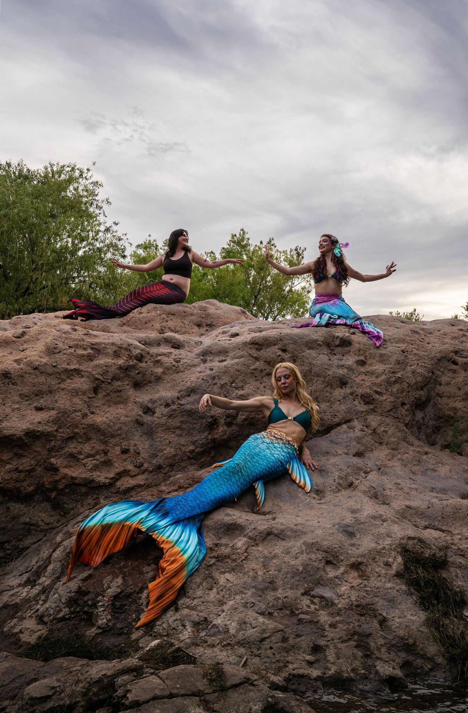 Three women in colorful mermaid costumes pose dramatically on large rocks near a river. Fantasy Photoshoot