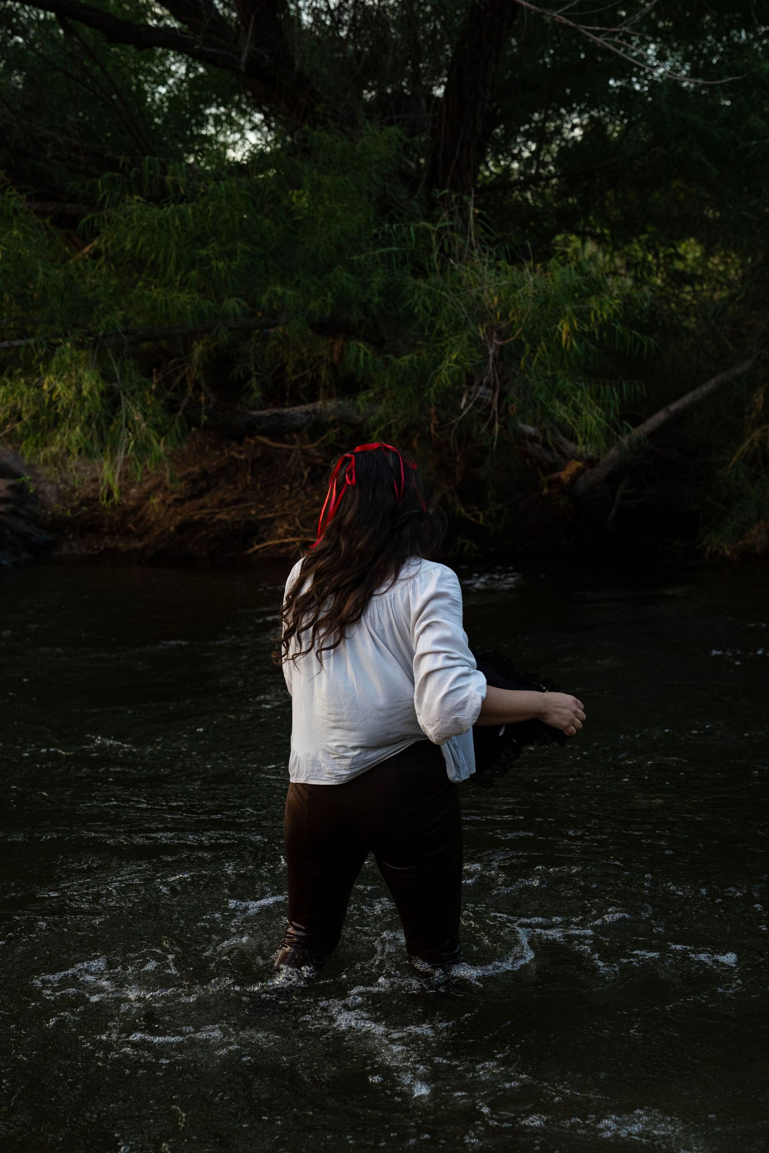 A woman in a white shirt and dark pants wades through a flowing river surrounded by dense green trees. Fantasy photoshoot