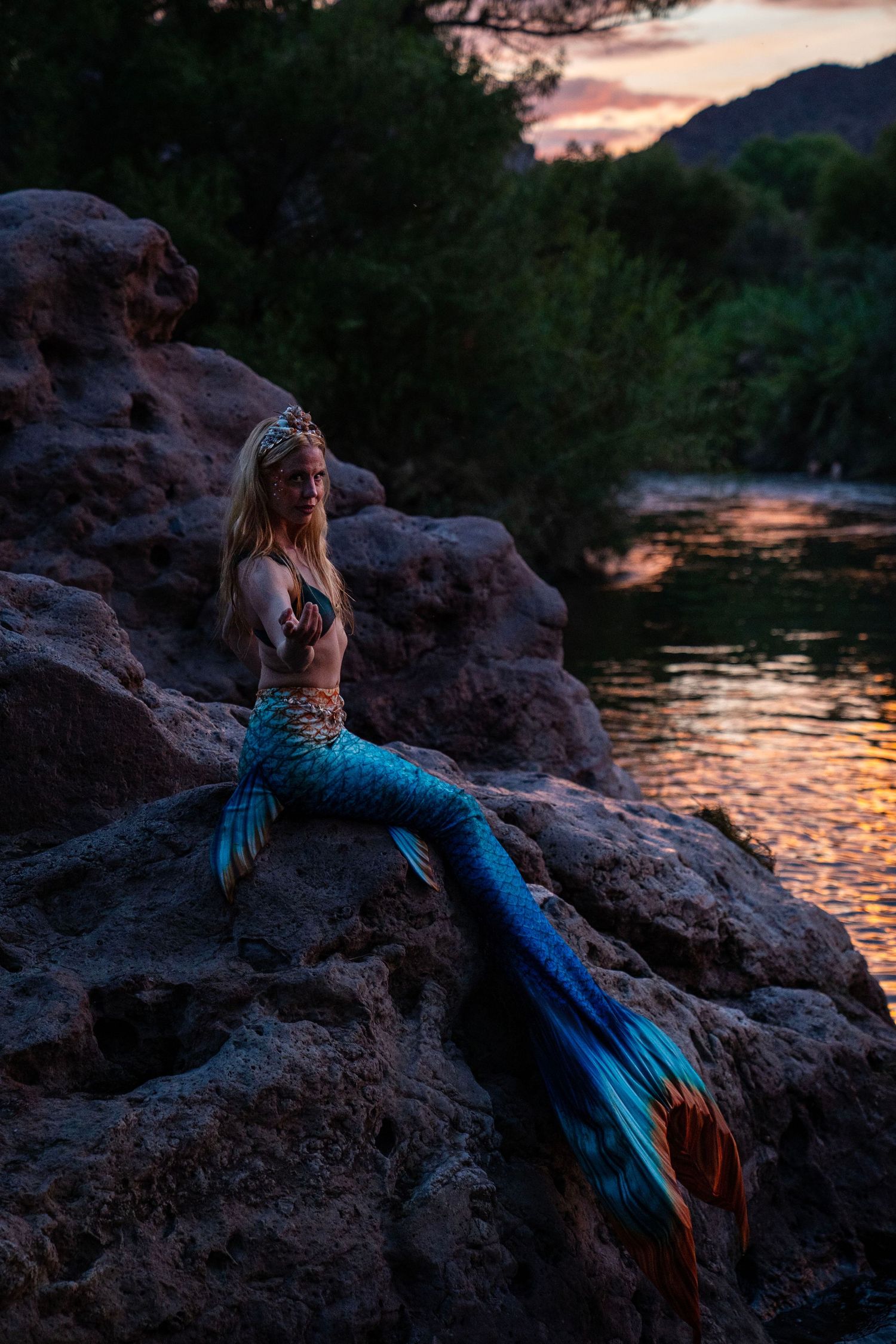 Blonde woman in blue mermaid tail and crown posing on rocks at sunset. Mermaid photoshoot, fantasy photoshoot. Arizona photographer.