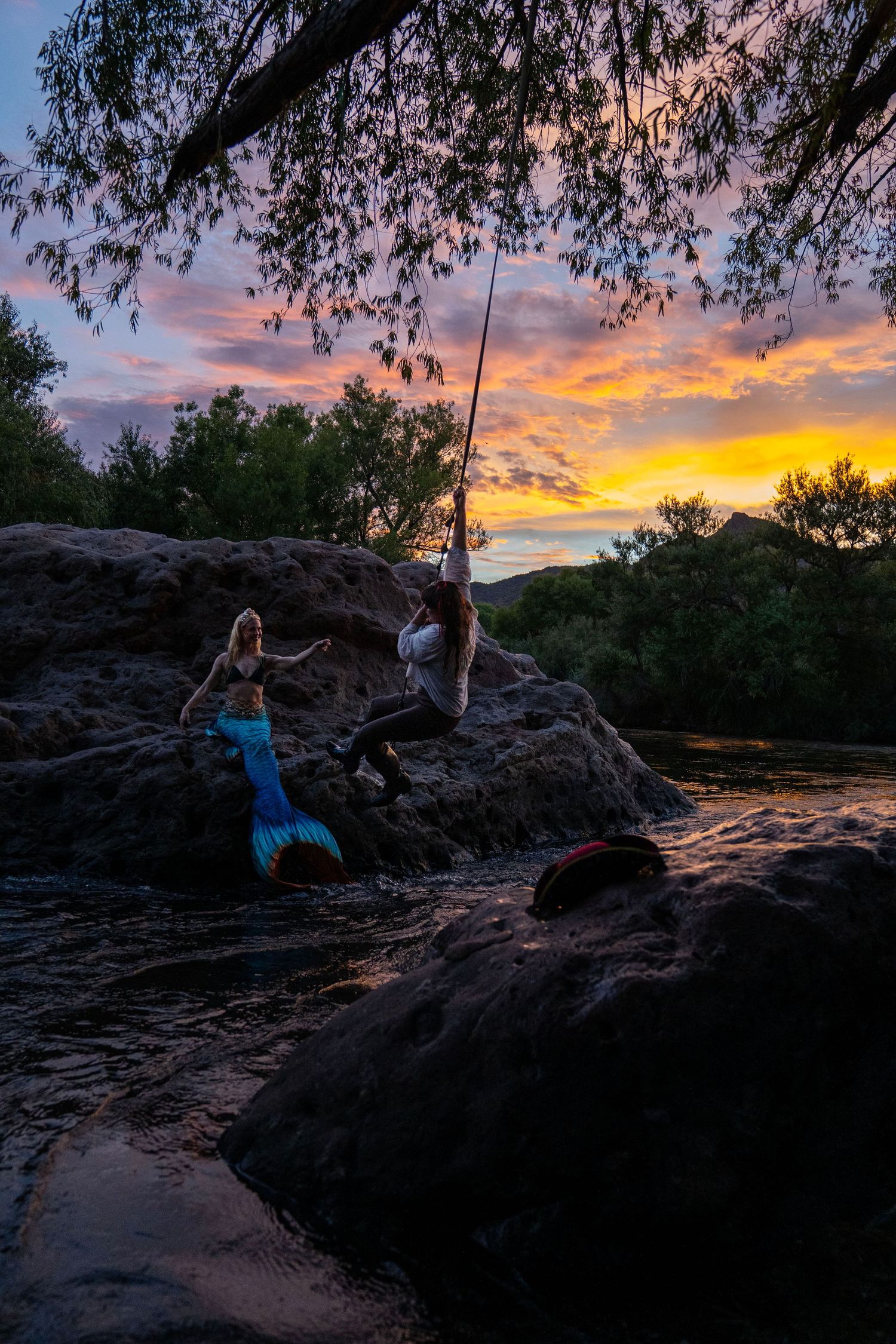 Mermaid and pirate swinging on rope over rocky river at sunset. Mermaid photoshoot, fantasy photoshoot. Arizona photographer.