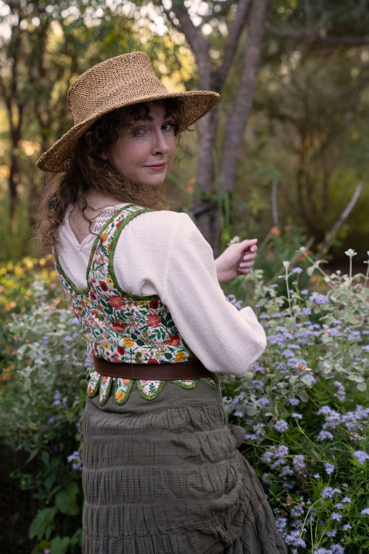 Woman in a straw hat and floral embroidered vest over a cream blouse and olive tiered skirt, standing among purple wildflowers outdoors.