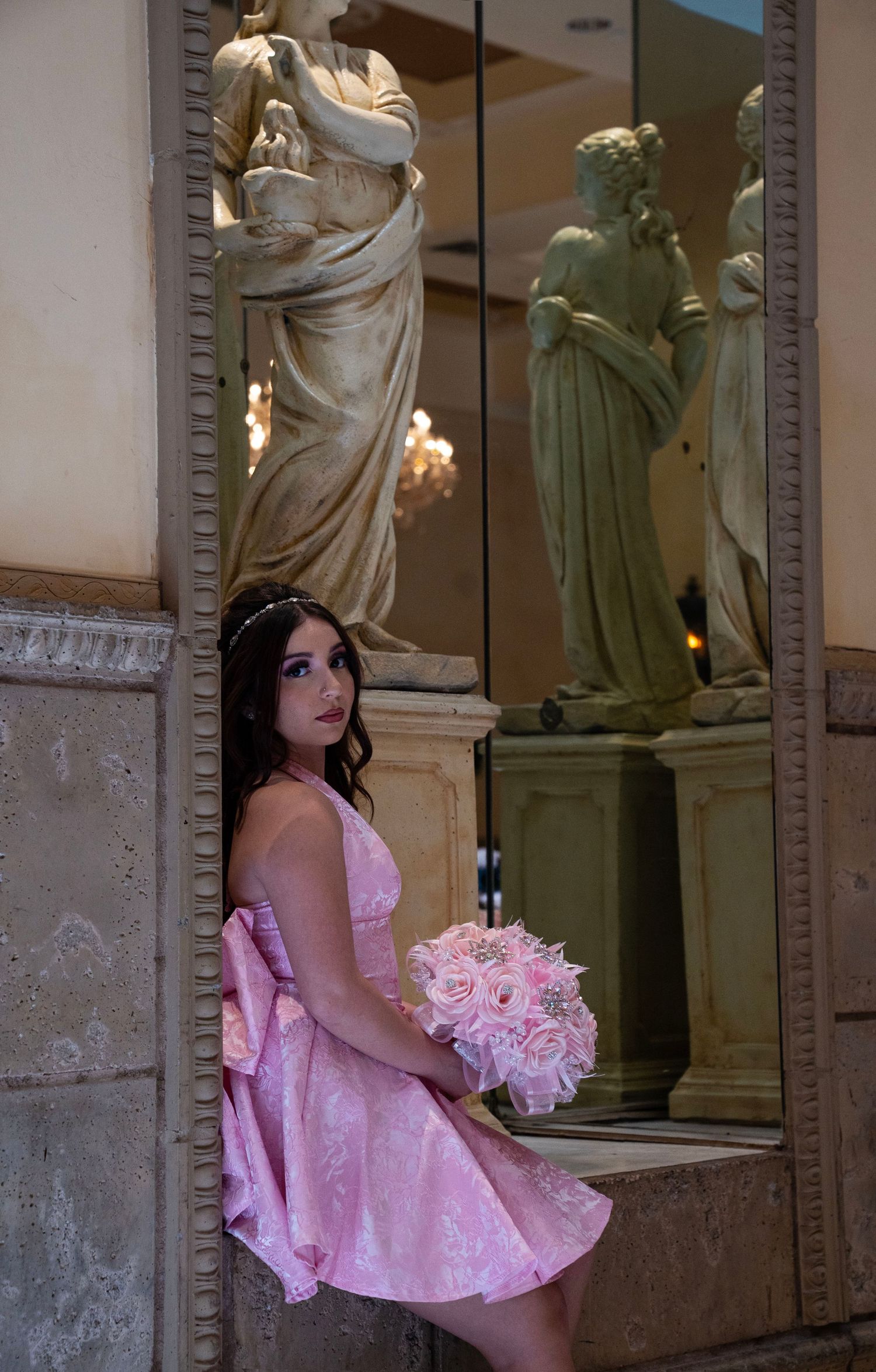 Elegant teen in pink dress holding bouquet beside classical statue in mirror.