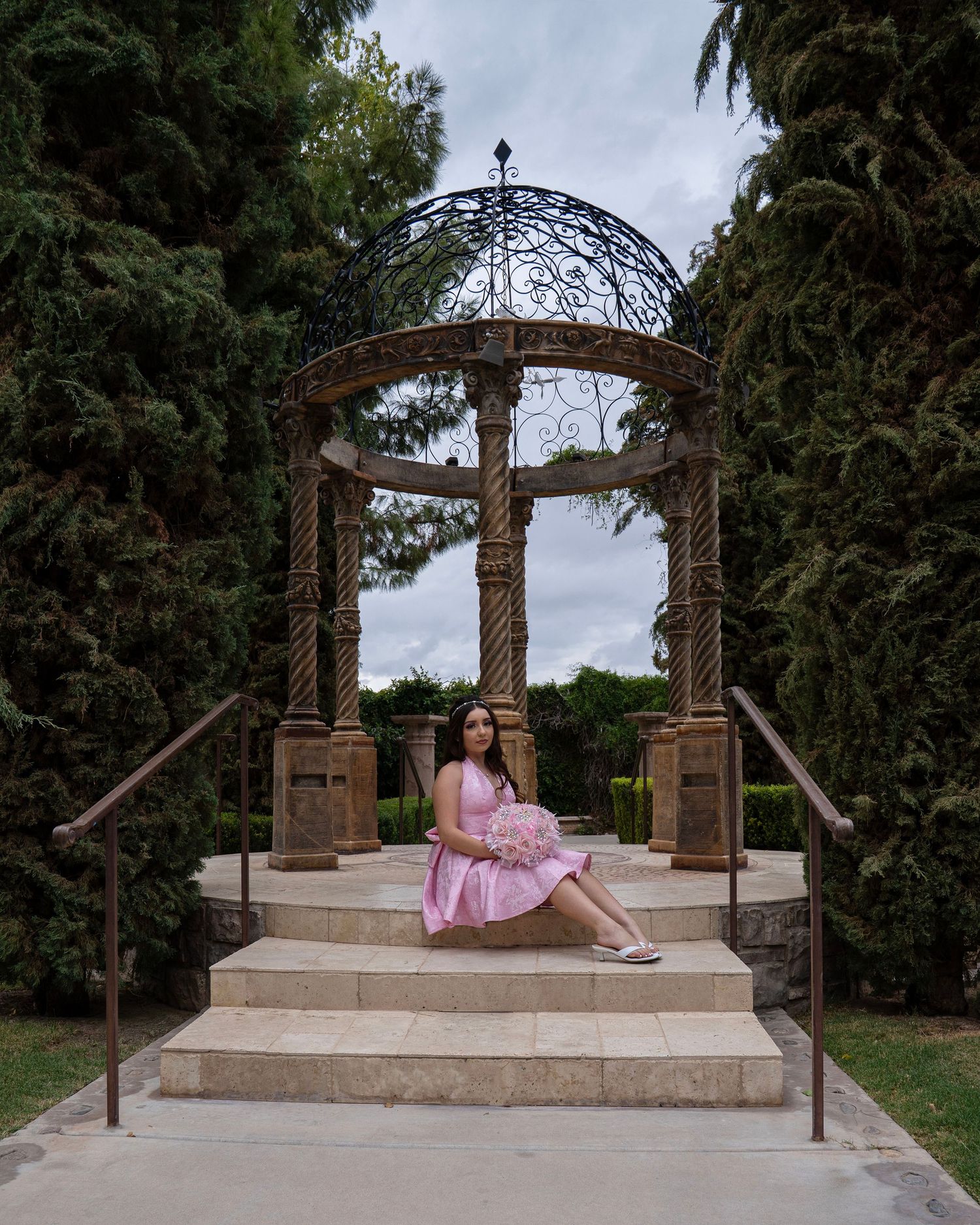 Young woman in pink dress sitting on gazebo steps surrounded by tall trees in an elegant garden setting.