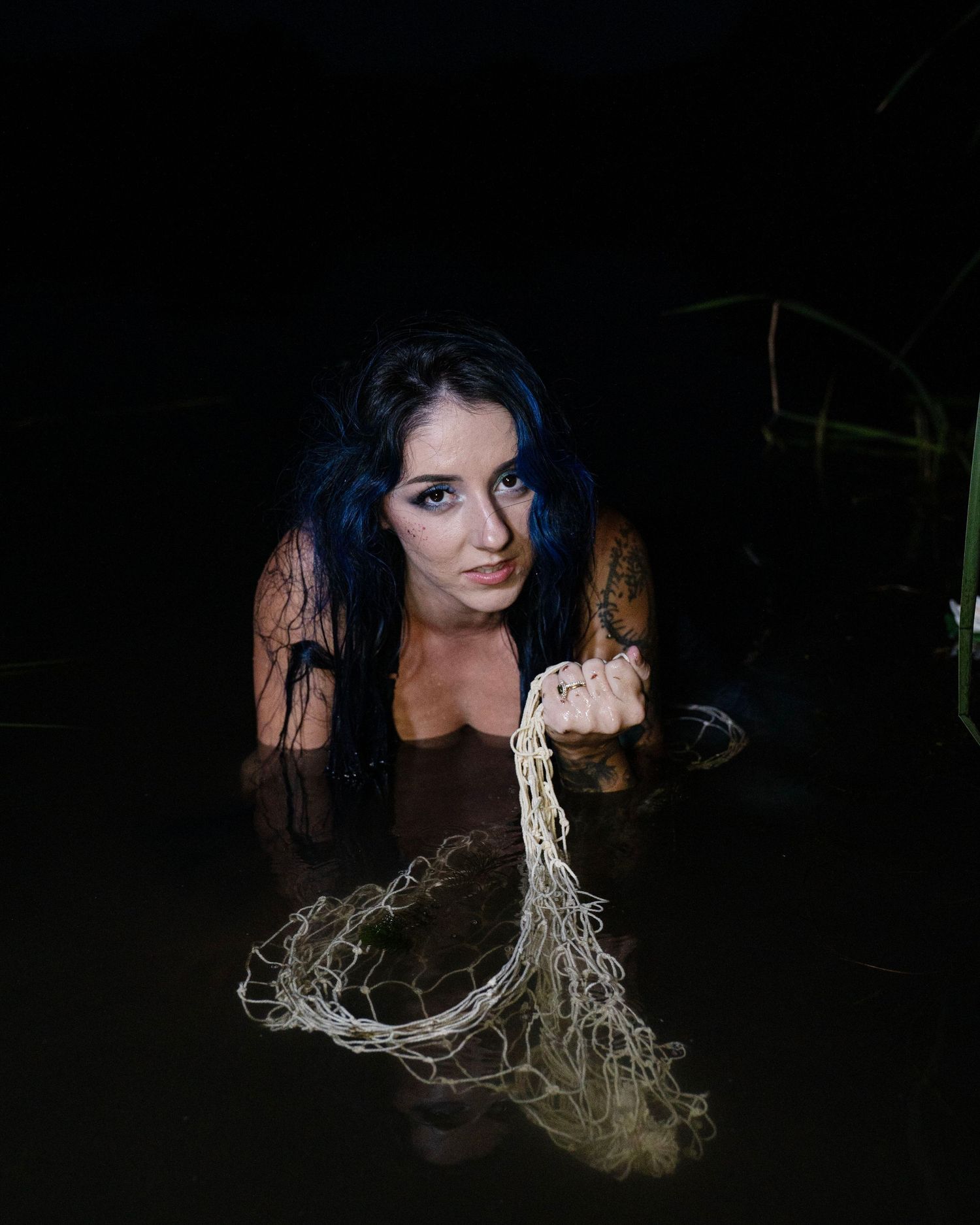 Woman with dark hair emerging from dark water, holding a rope net, with a mysterious and dramatic atmosphere.