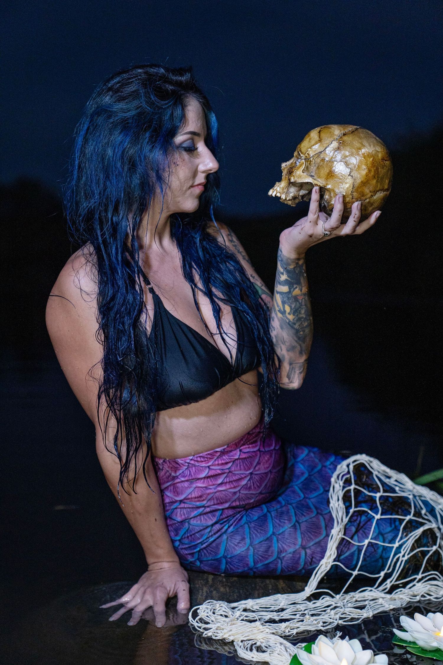 Tattooed woman with blue hair in mermaid costume holds a skull near water at night.