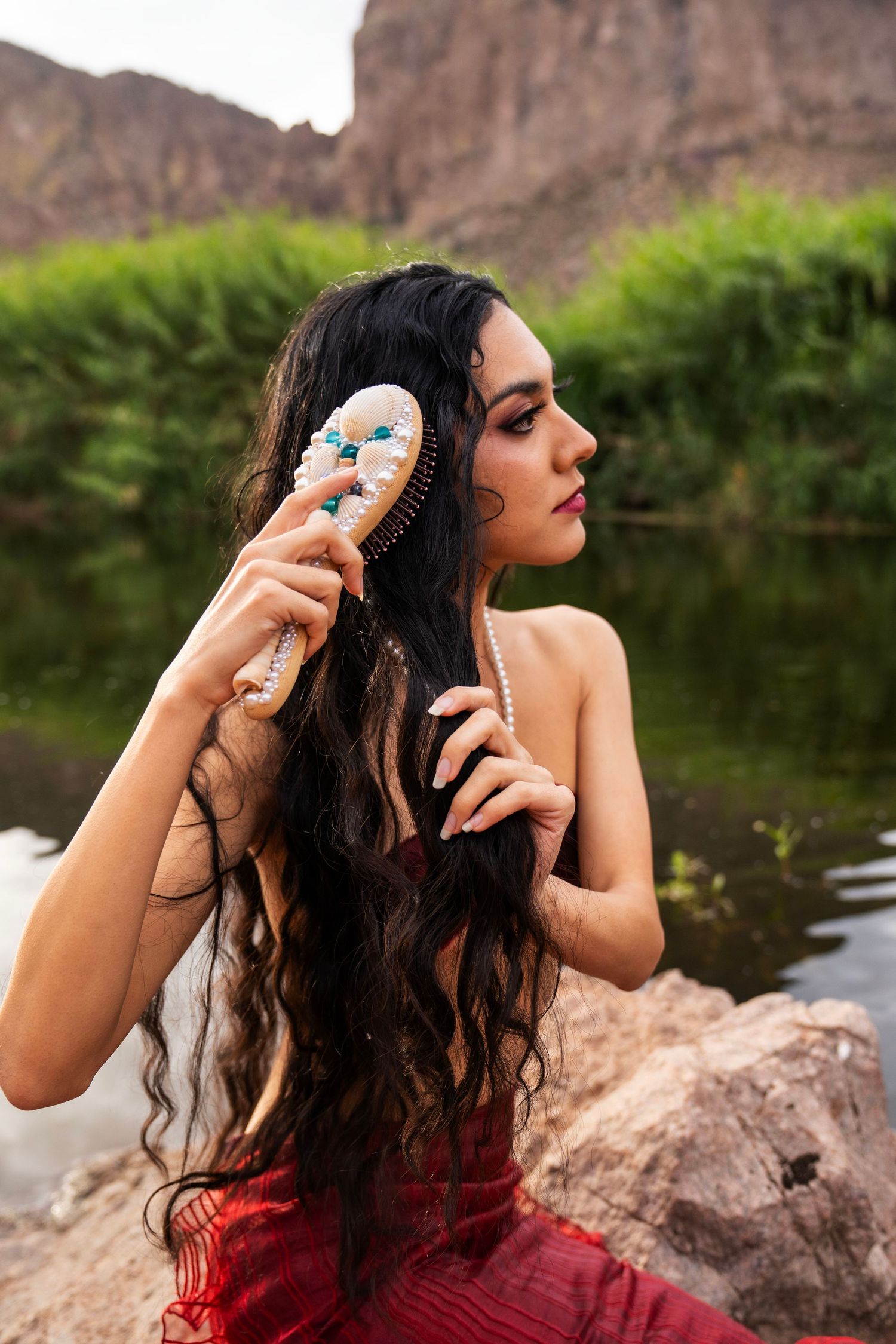 A dark-haired woman in a red mermaid outfit brushes her long wavy hair with a decorative brush by a river.