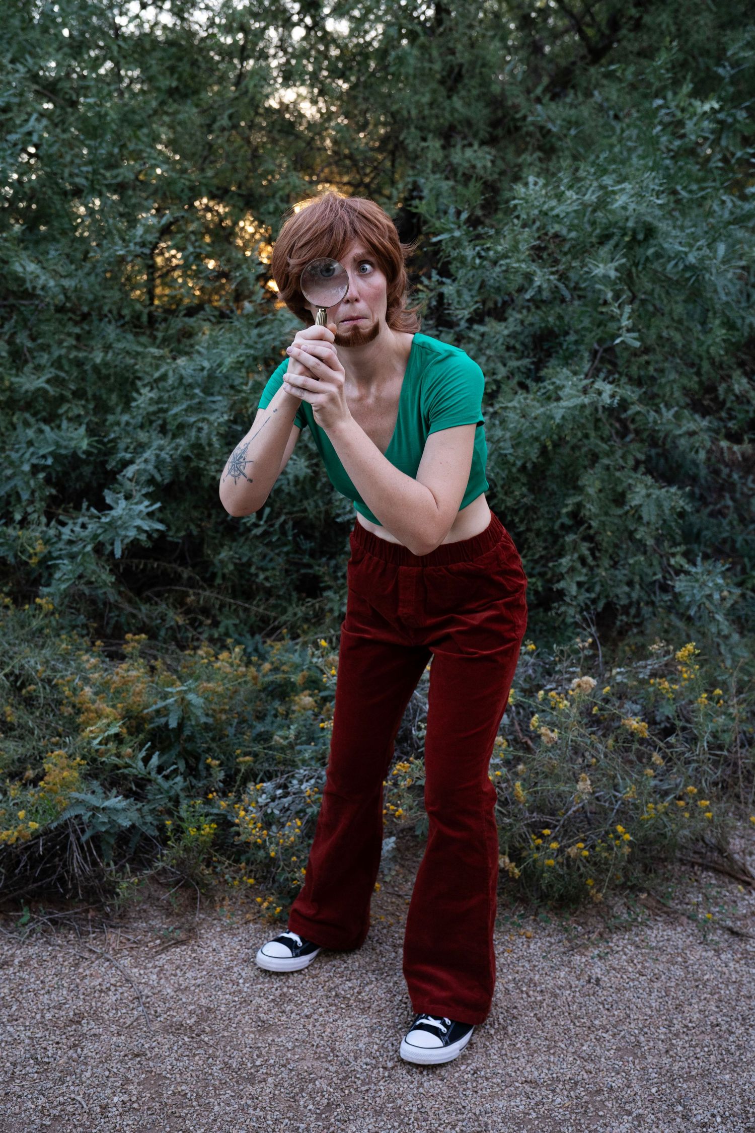 Person in a green top and red corduroy flared pants peers through a magnifying glass while standing outdoors among leafy green shrubs.