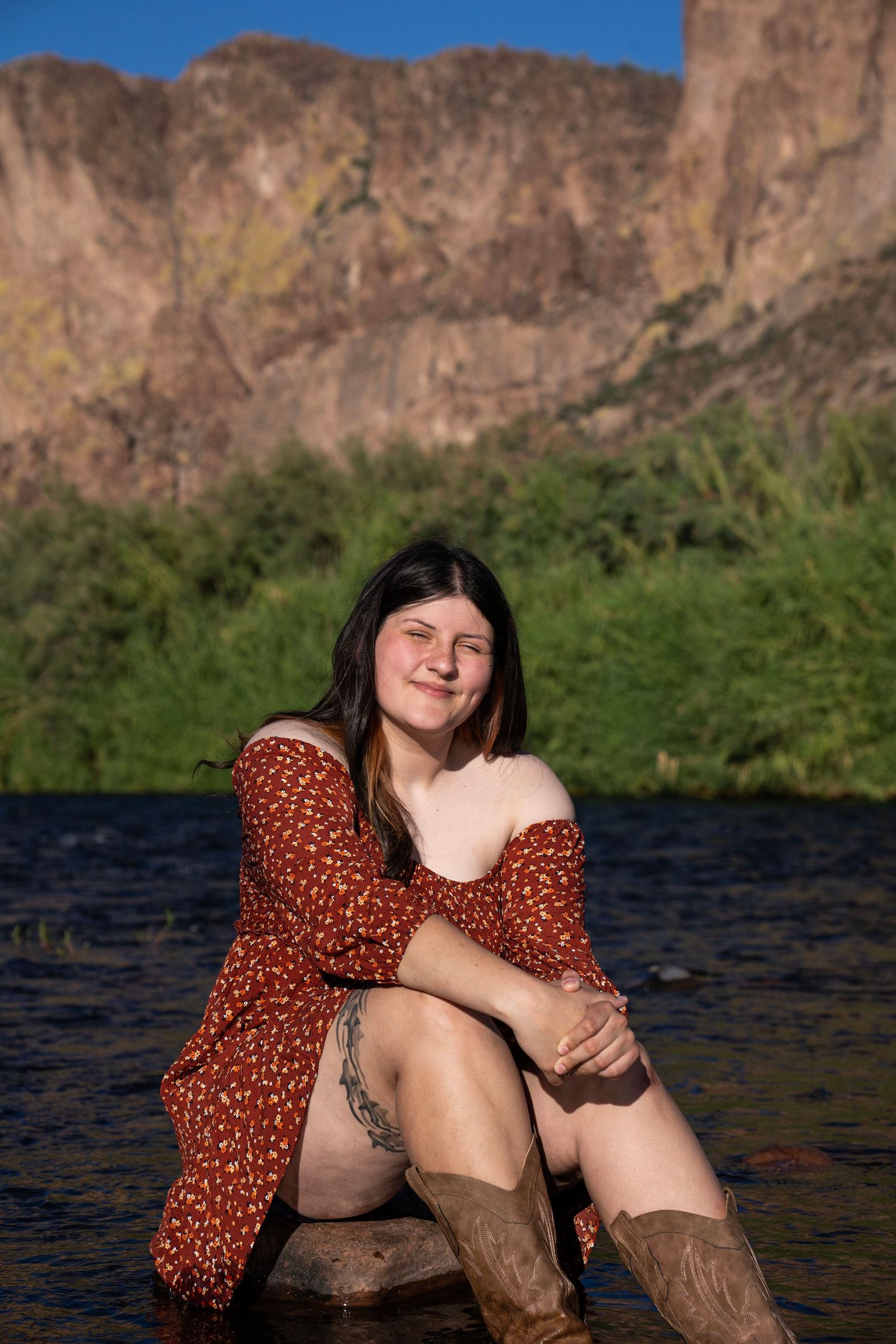 Young woman in red floral dress and cowboy boots sitting by a river with rocky mountain cliffs in background.