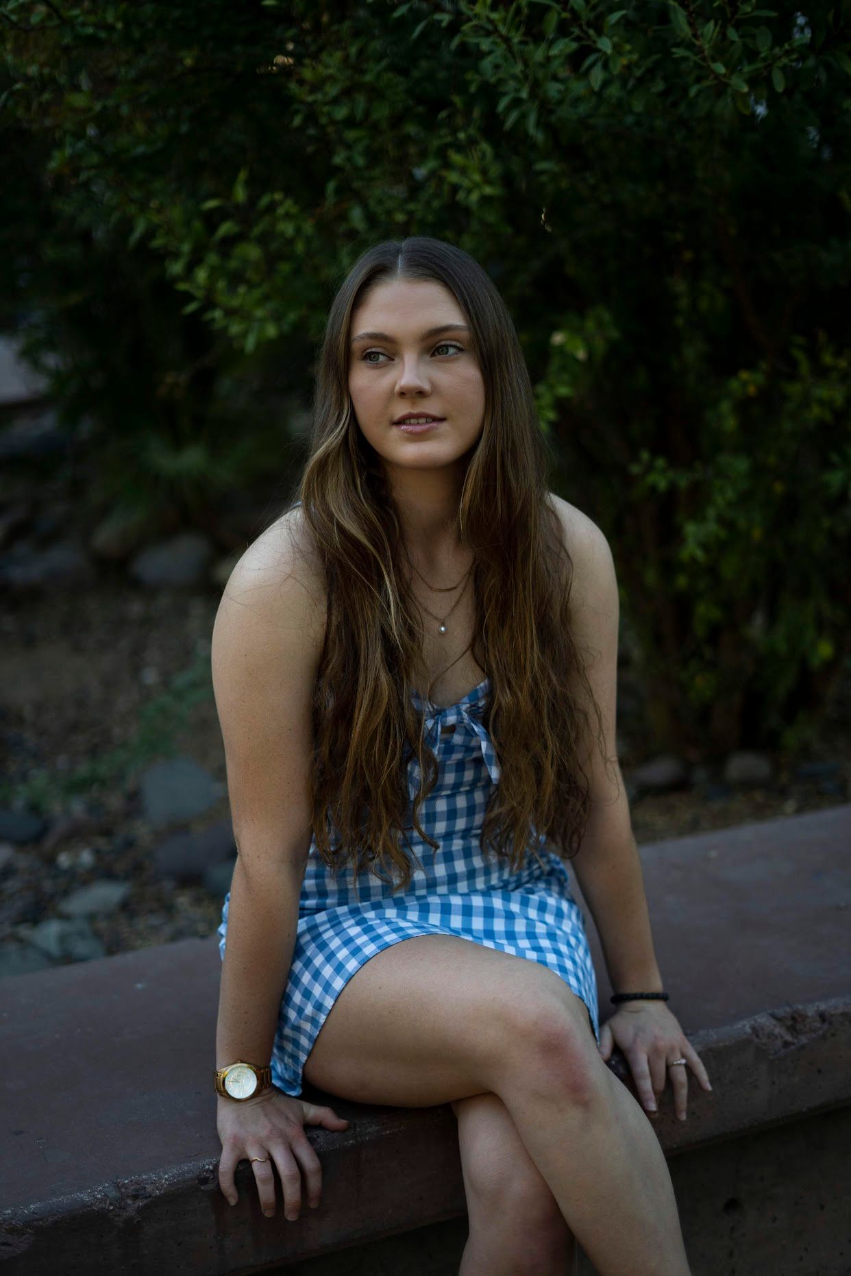 Young woman in blue gingham dress sitting on bench outdoors.