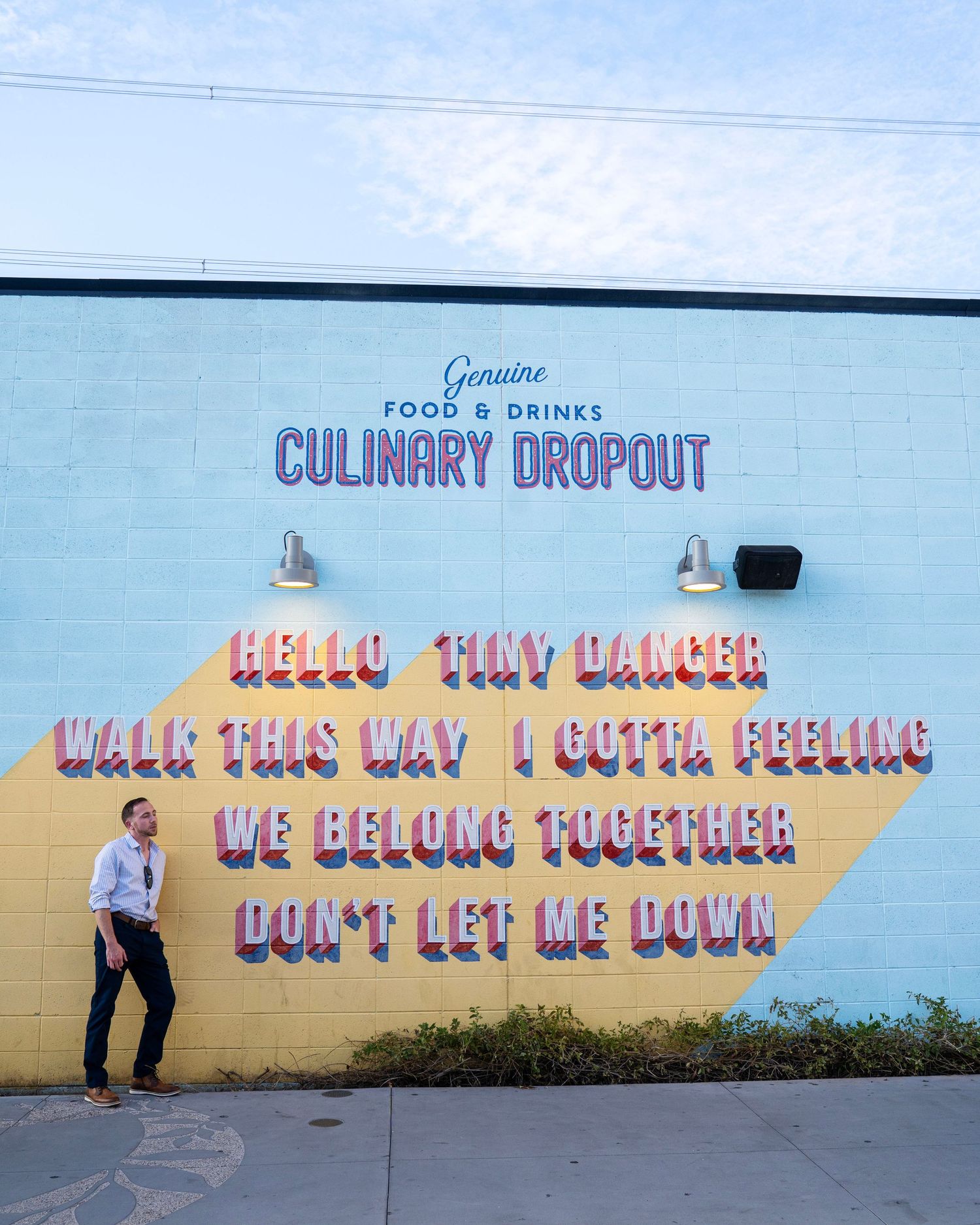 Colorful Culinary Dropout restaurant mural with song lyrics on a light blue and yellow wall with a man walking by.