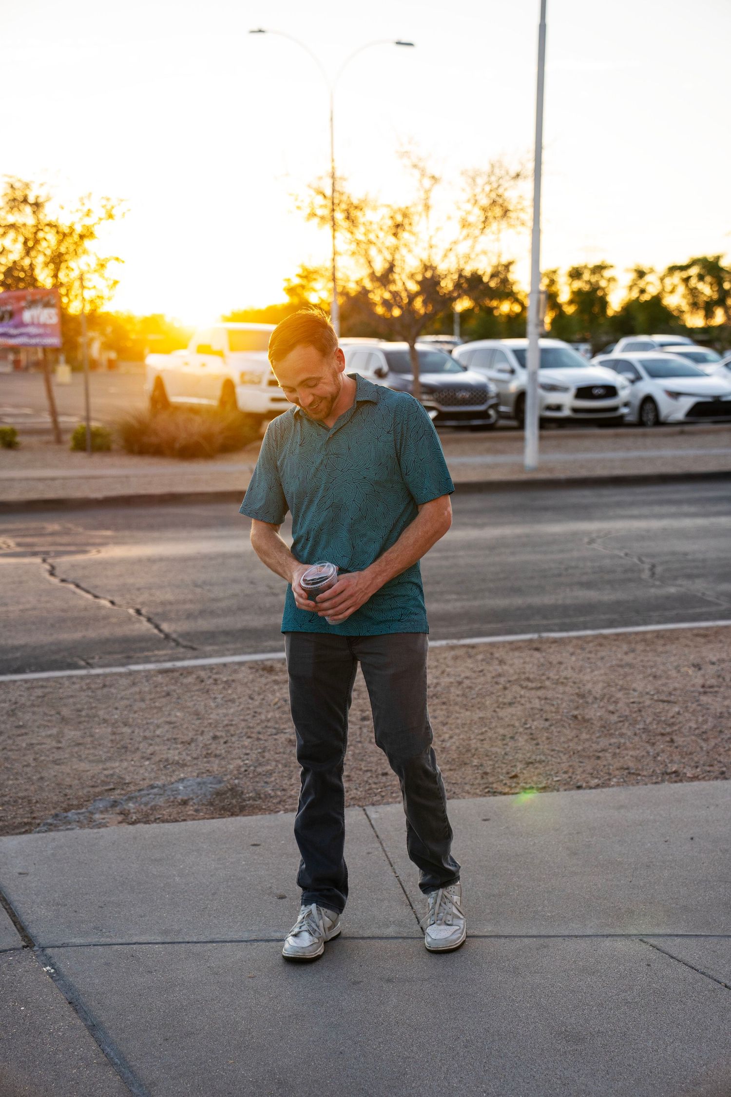 Man in teal polo shirt checking phone on sidewalk during golden hour sunset with parking lot in background.