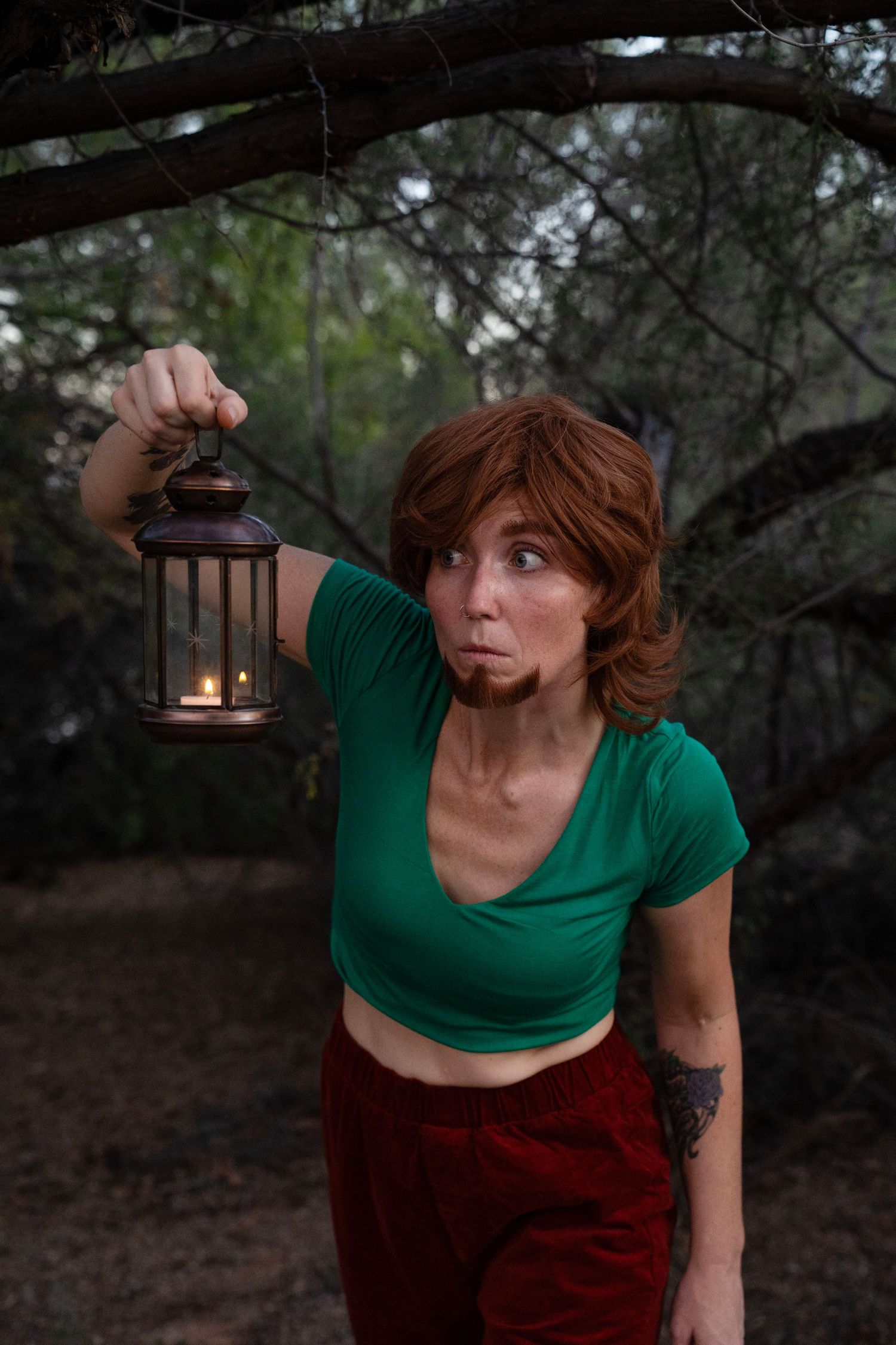 Woman in a green crop top and red pants holds a lit vintage lantern while peering cautiously through a dim, wooded forest.