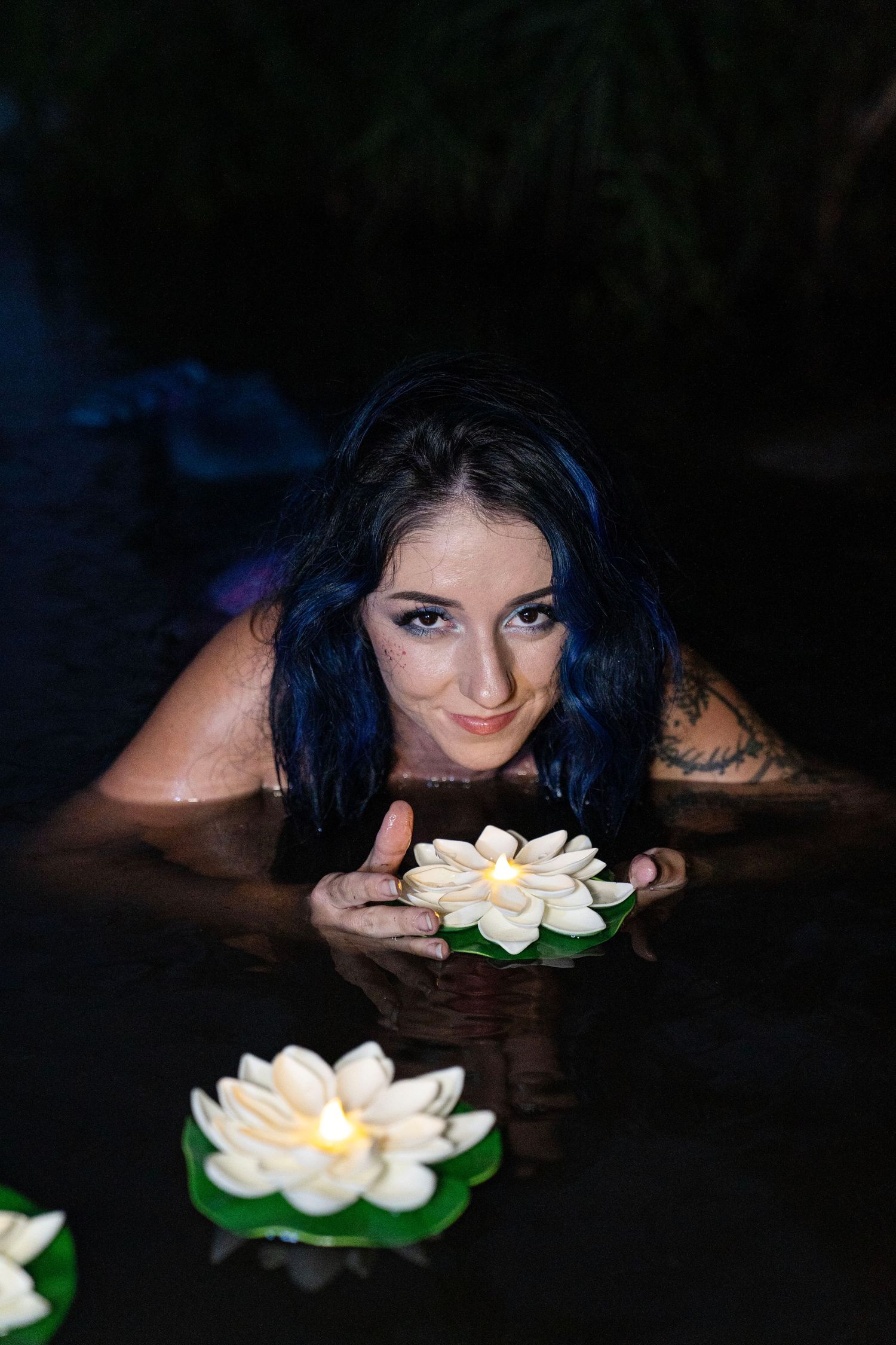 Blue-haired woman with tattoos poses in dark water with floating white lotus flowers.