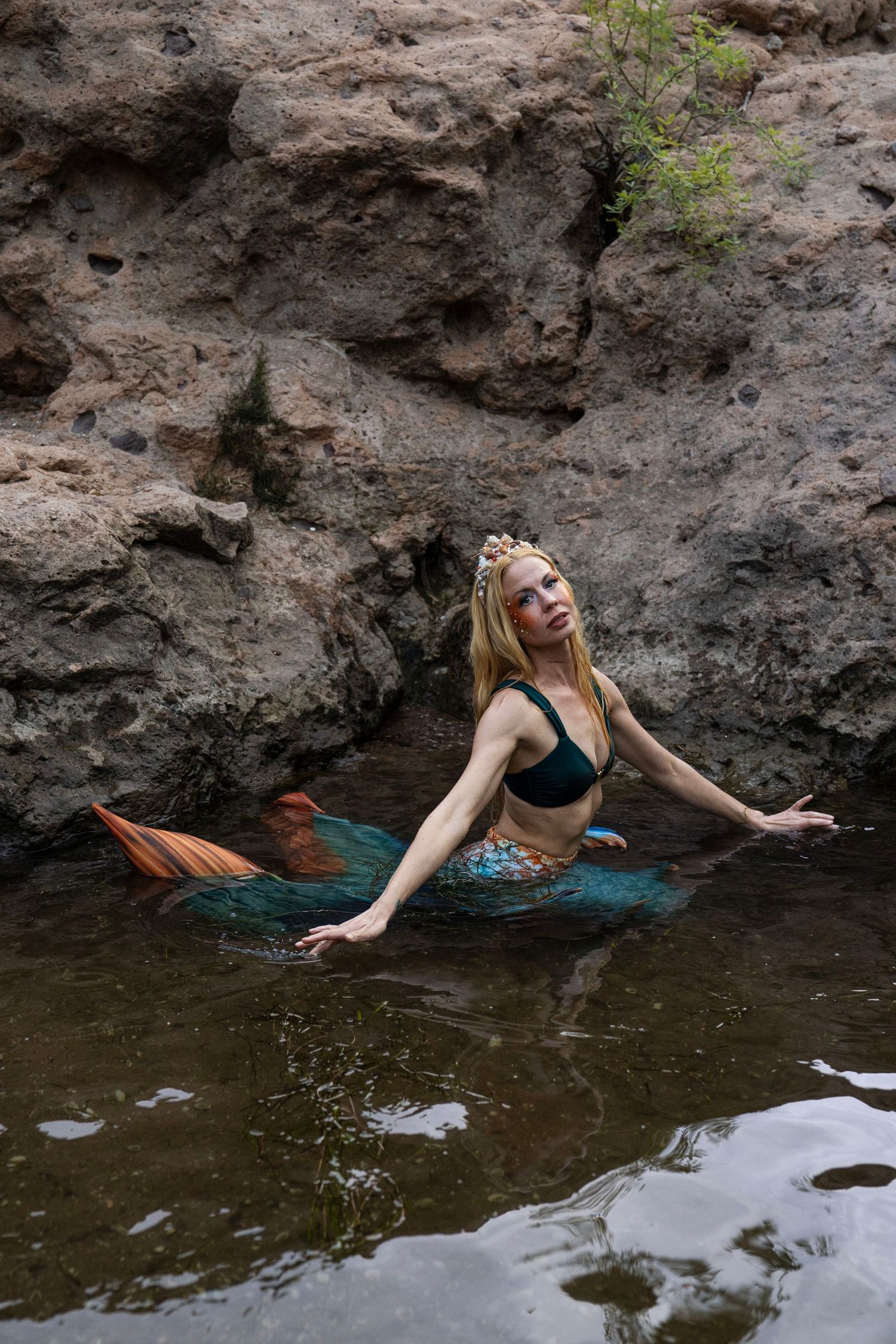 Blonde woman wearing mermaid tail and crown gazing upward while posing in natural rocky water. Fantasy Photoshoot