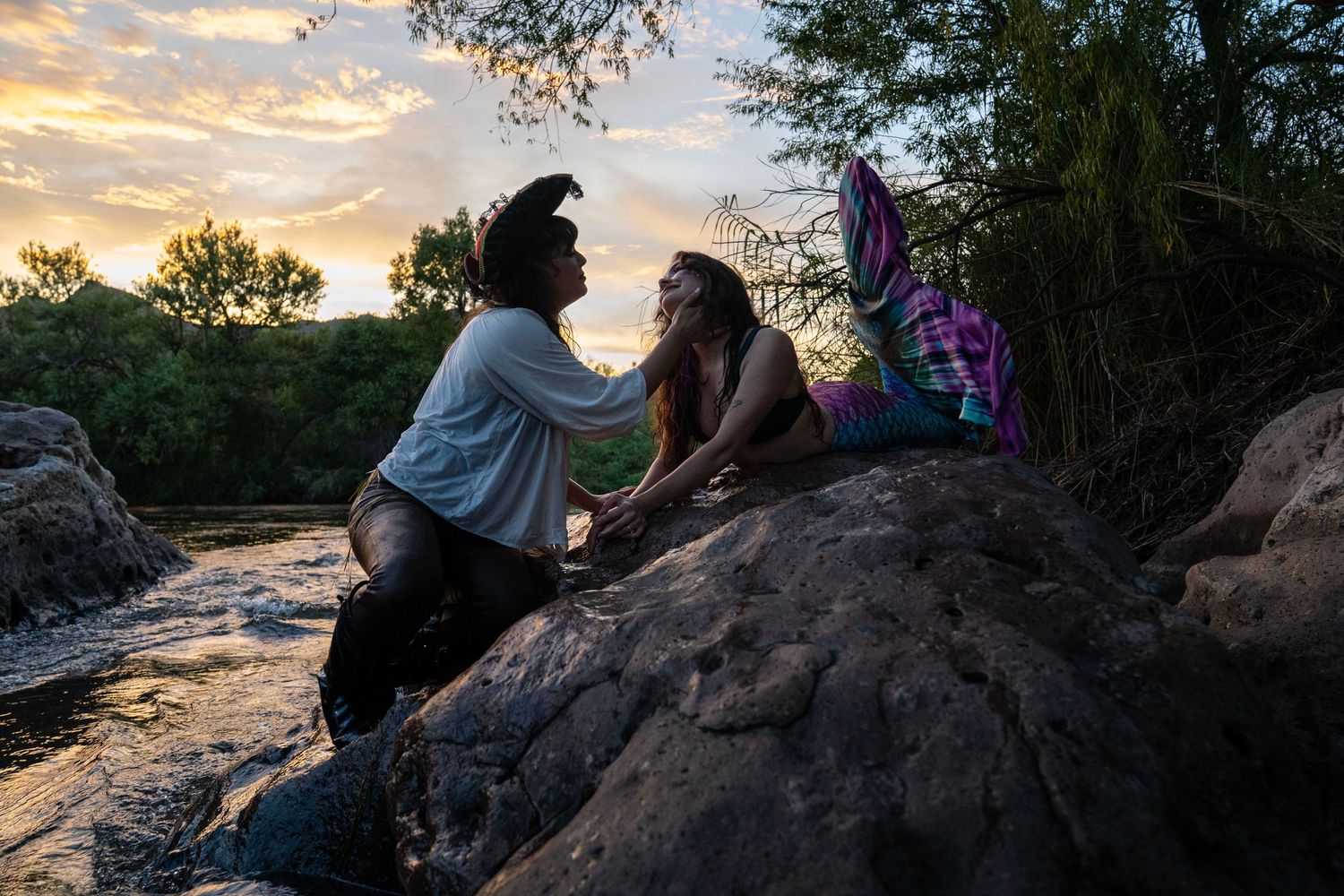 Pirate and mermaid costume models sitting on riverside rocks during a colorful sunset. Mermaid photoshoot, fantasy photoshoot.