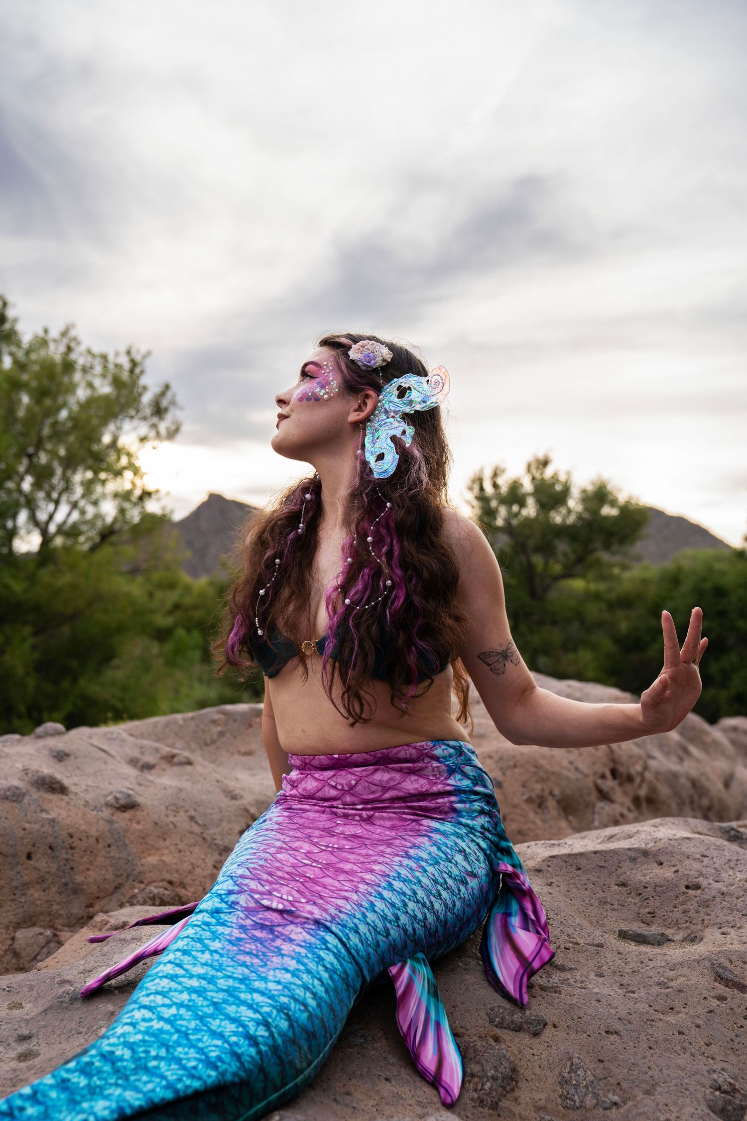 Woman in a pink and teal mermaid tail with elaborate face paint and hair accessories posing on rocks. Fantasy Photoshoot